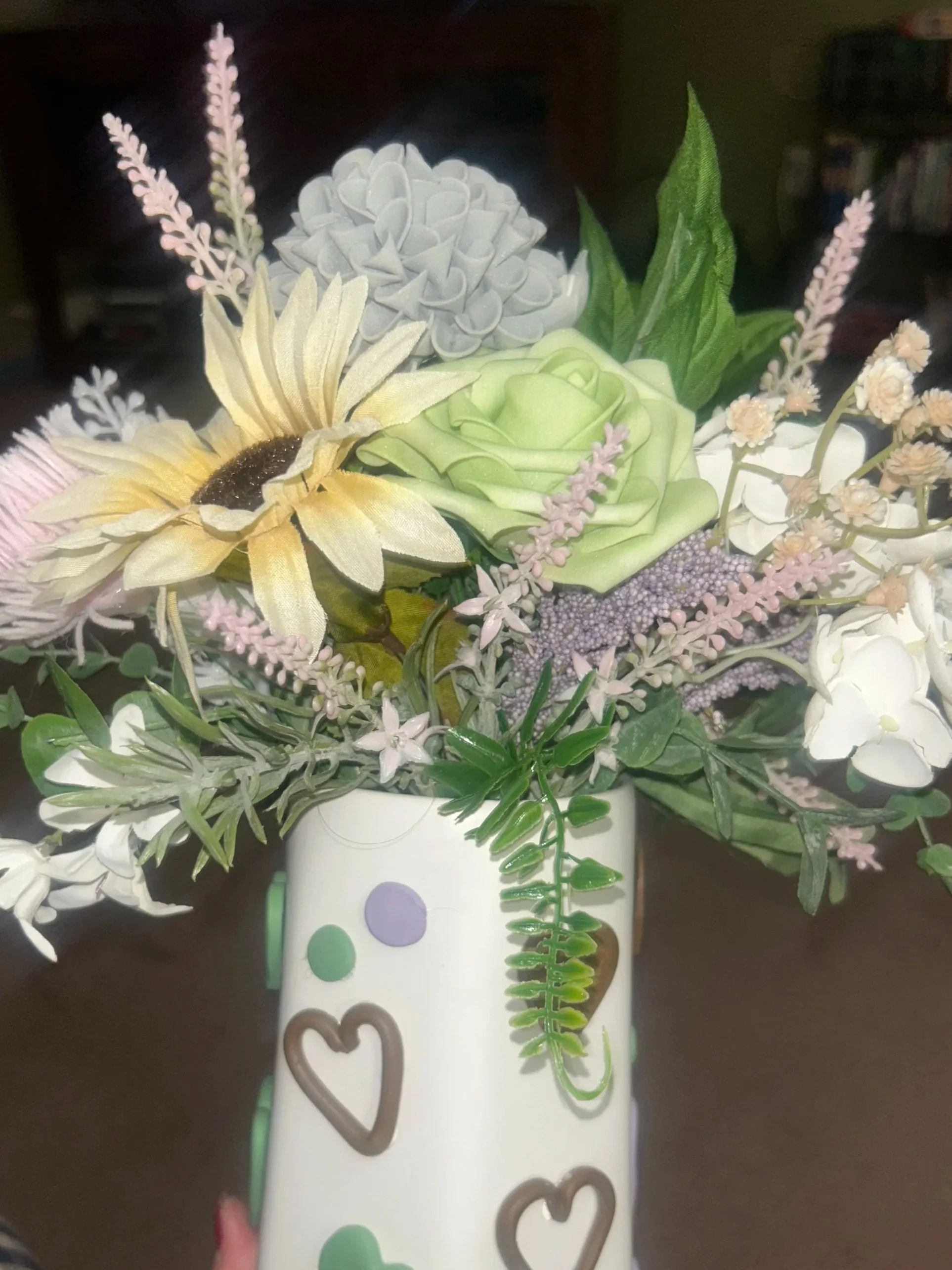 A bouquet of artificial flowers in a white vase with green and purple polka dots, adorned with a decorative heart-shaped wire, includes a yellow sunflower, a green rose, lavender sprigs, and various greenery.