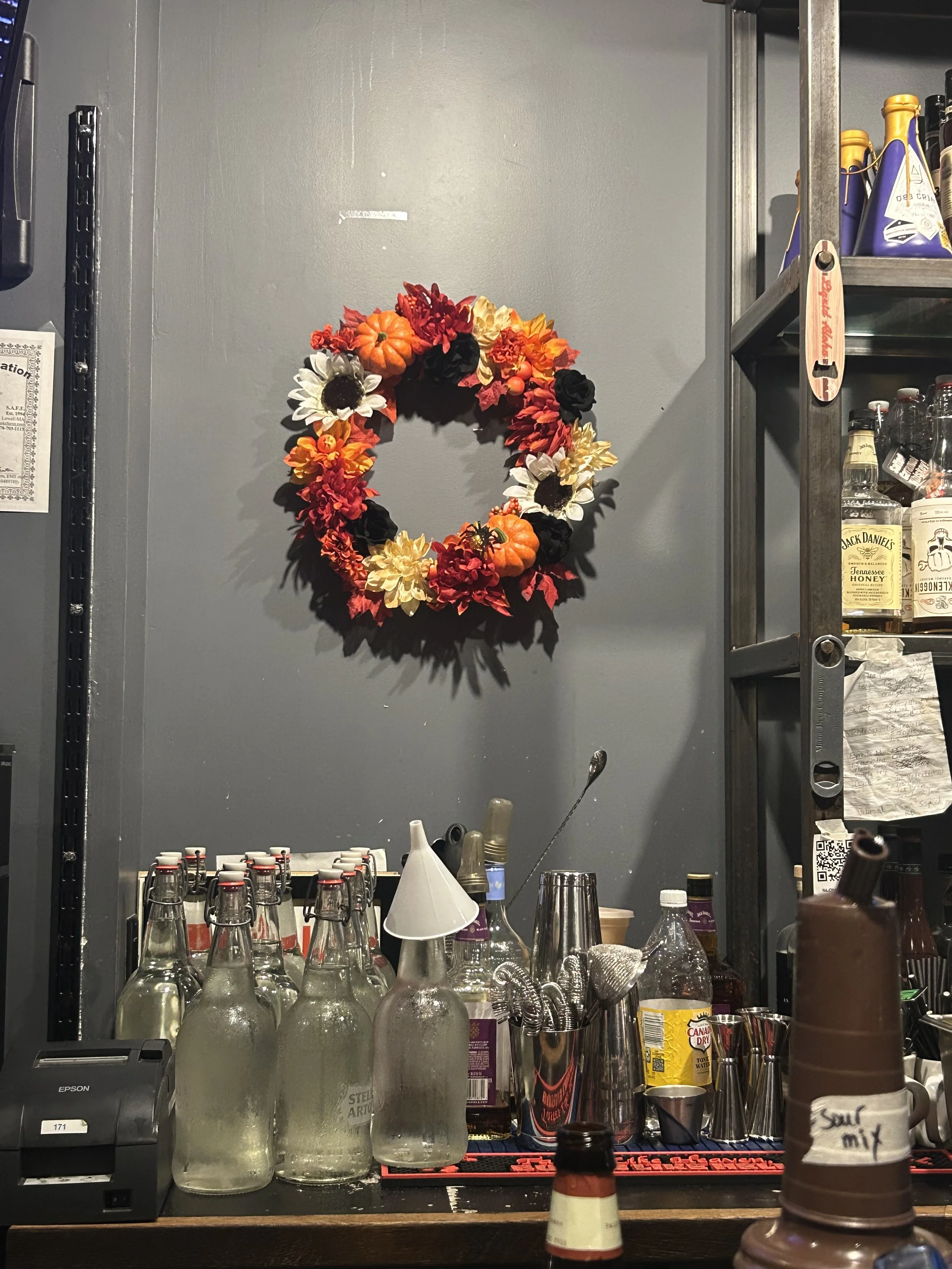 Shelf with decorative fall wreath, various bottles and glasses, and bar tools.