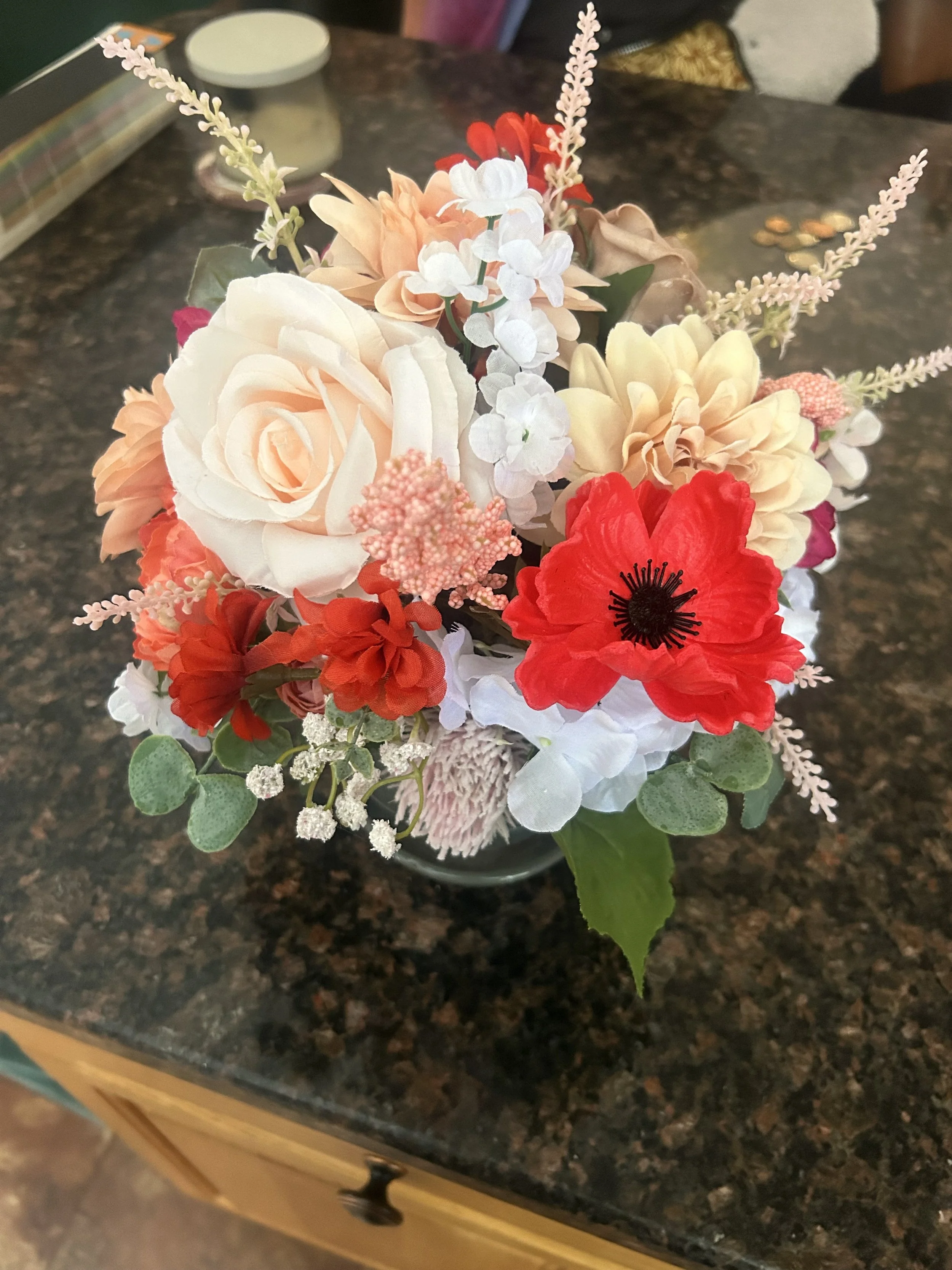 A bouquet of artificial flowers, including white, red, and peach-colored roses, along with small white and pink blossoms, placed on a dark granite kitchen counter.