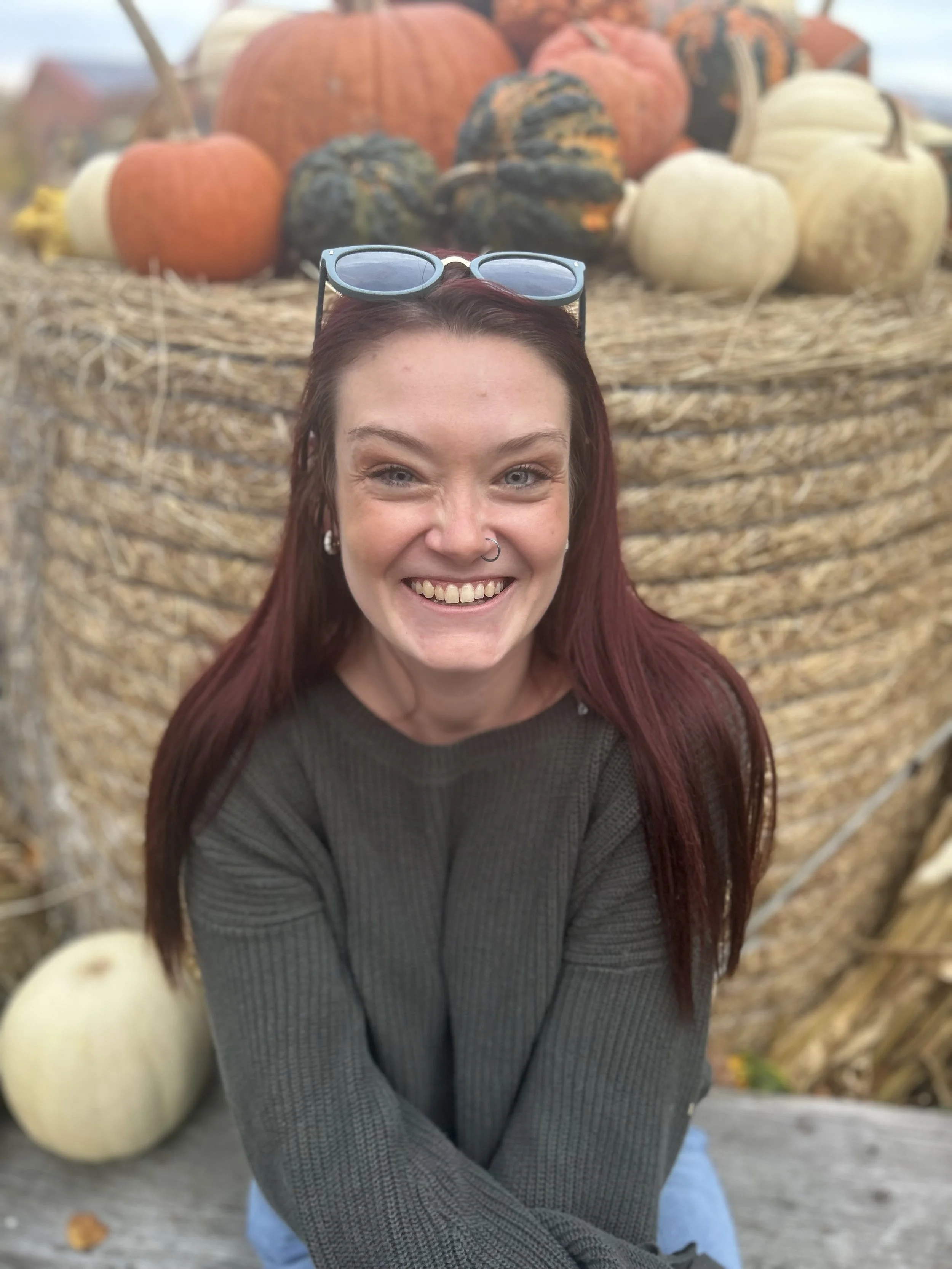 A smiling woman with red hair and sunglasses on her head taking a selfie in front of a display of pumpkins.