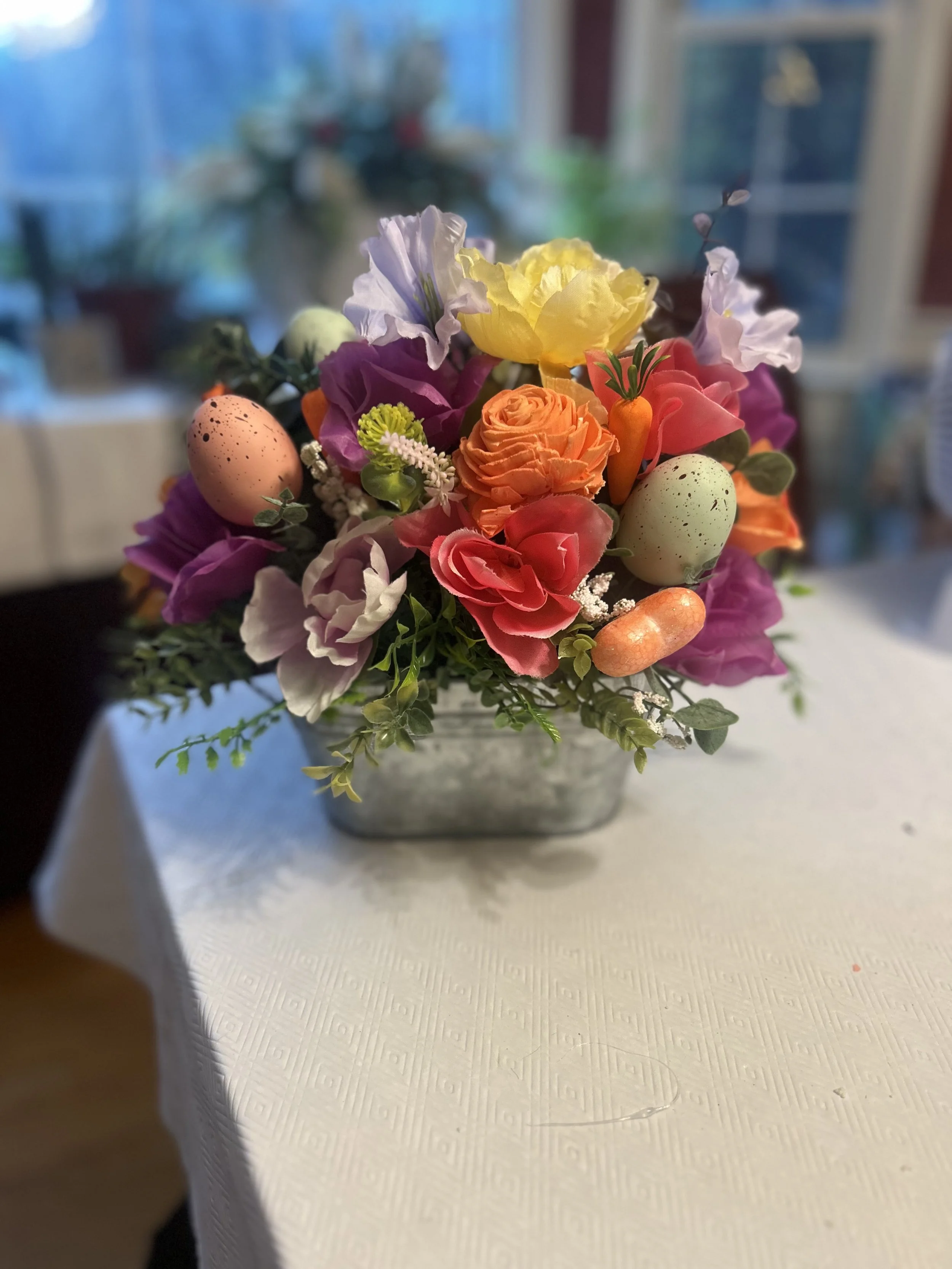 A floral arrangement in a metal container with various colorful flowers and decorative eggs on a white tablecloth.
