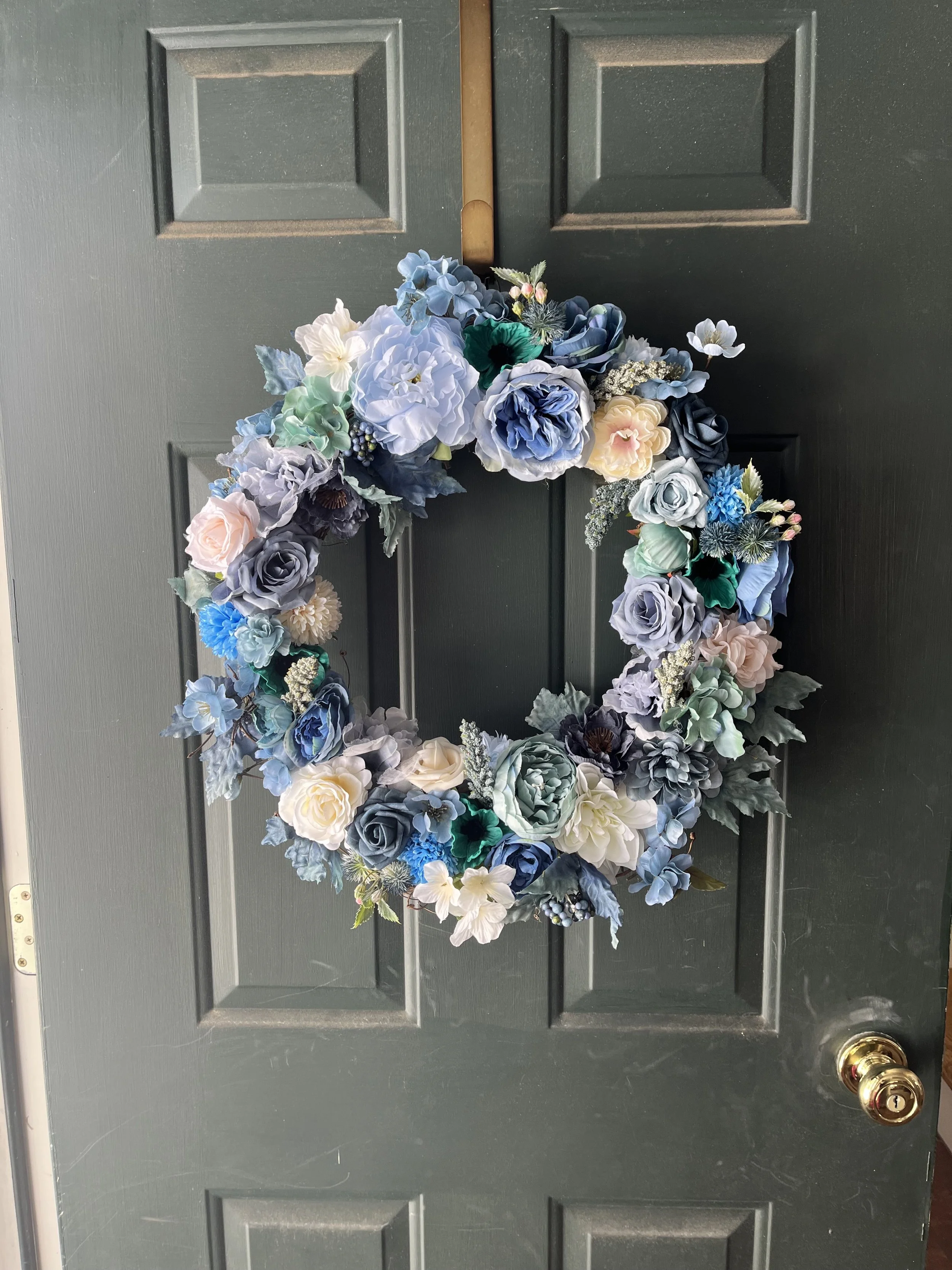 A decorative wreath made of blue, white, and pale yellow artificial flowers hanging on a dark green door with a gold doorknob.