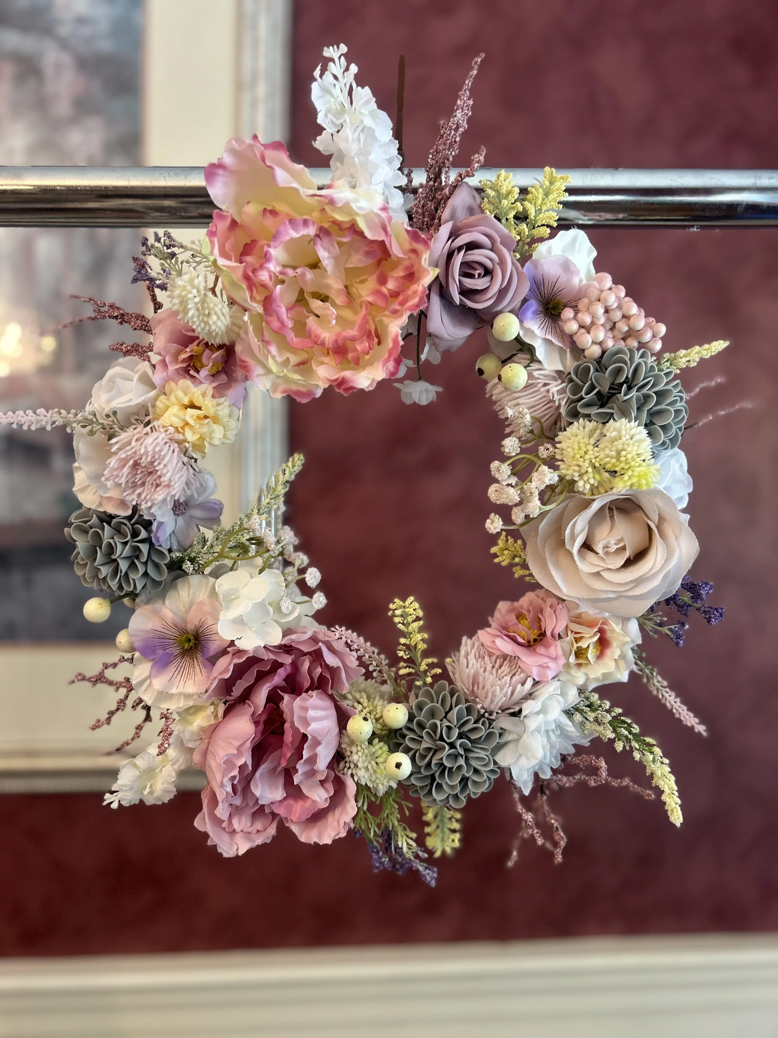 Colorful flower wreath with roses, hydrangeas, and greenery on a silver horizontal rod, against a blurred red background.