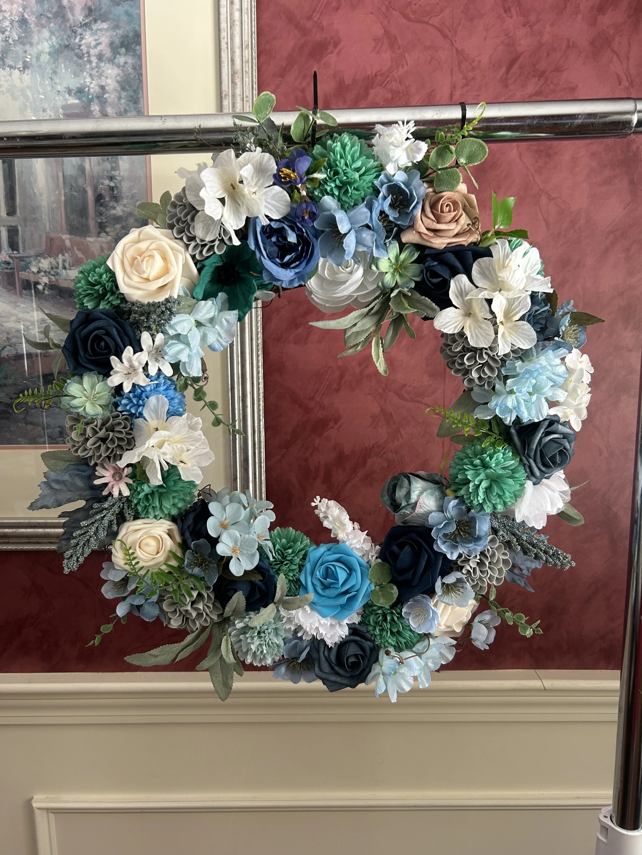Colorful floral arrangement with various artificial flowers including roses, hydrangeas, and other blooms, hanging on a mirror in front of a red textured wall.