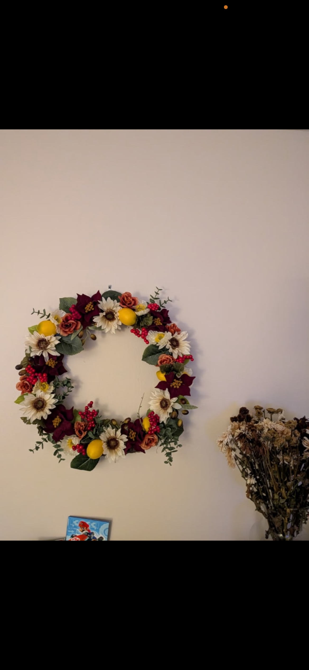 Decorative floral wreath with white, yellow, and red flowers hanging on a white wall, next to a bunch of dried flowers.