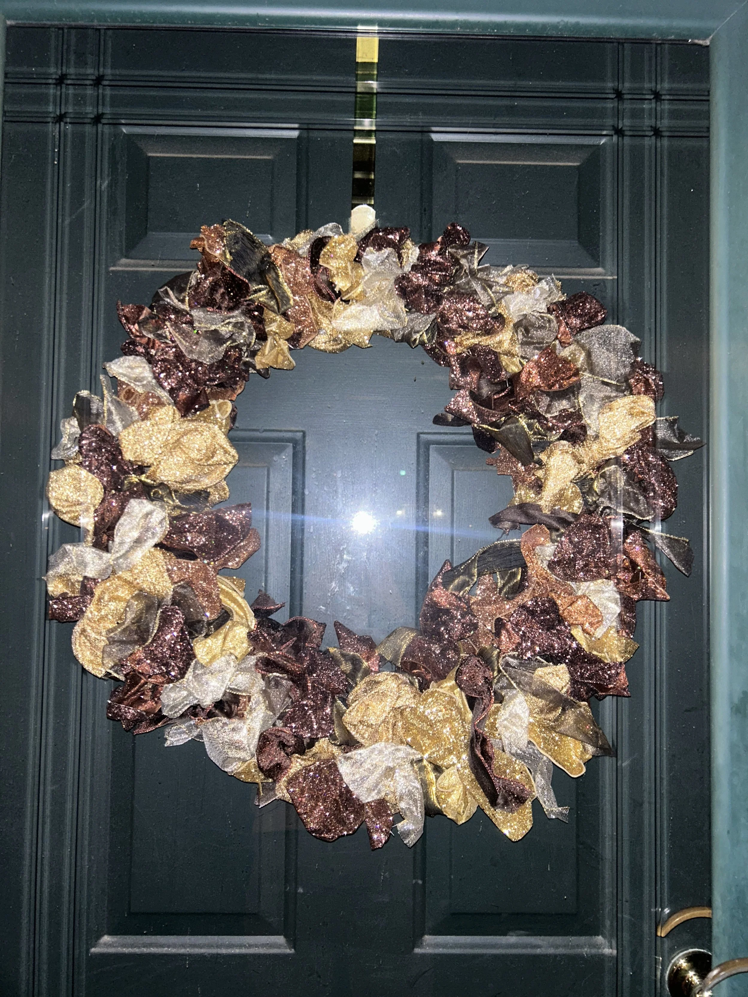 A Christmas wreath decorated with glittery ribbons in shades of gold, bronze, and black, hanging on a dark-colored door.