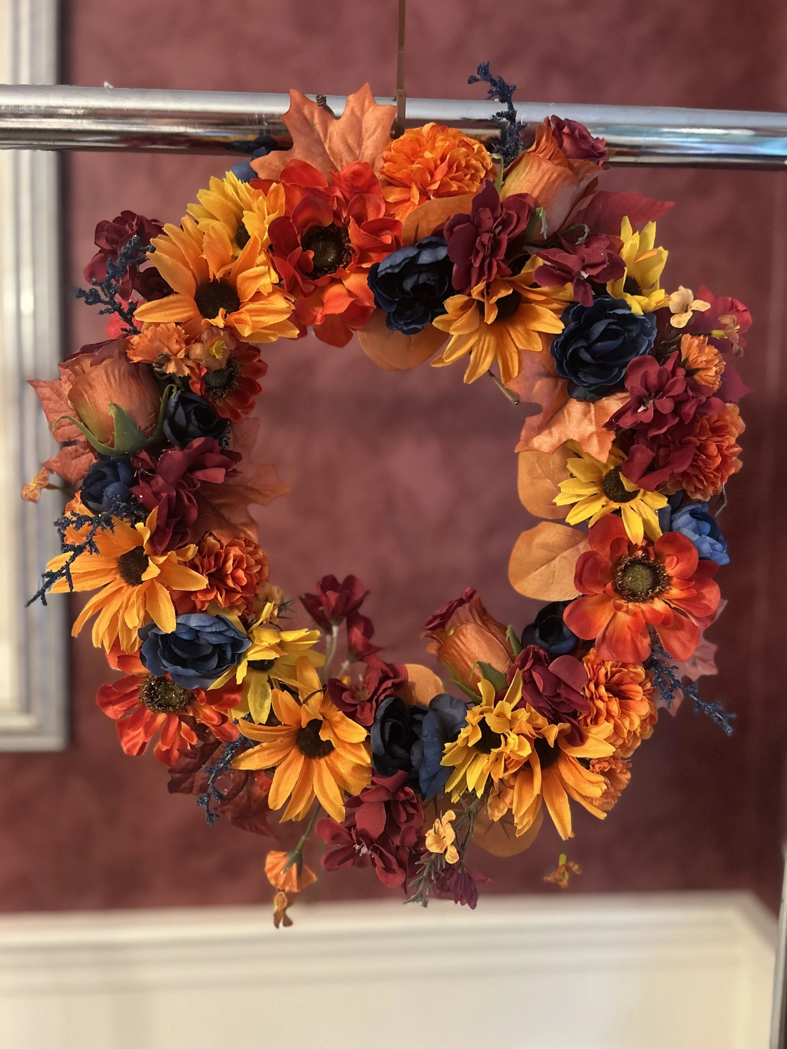 A circular autumn-themed flower wreath composed of orange, yellow, red, and dark blue flowers, with some leaves and sprigs of decorative foliage, hanging from a metal holder against a maroon carpeted background.