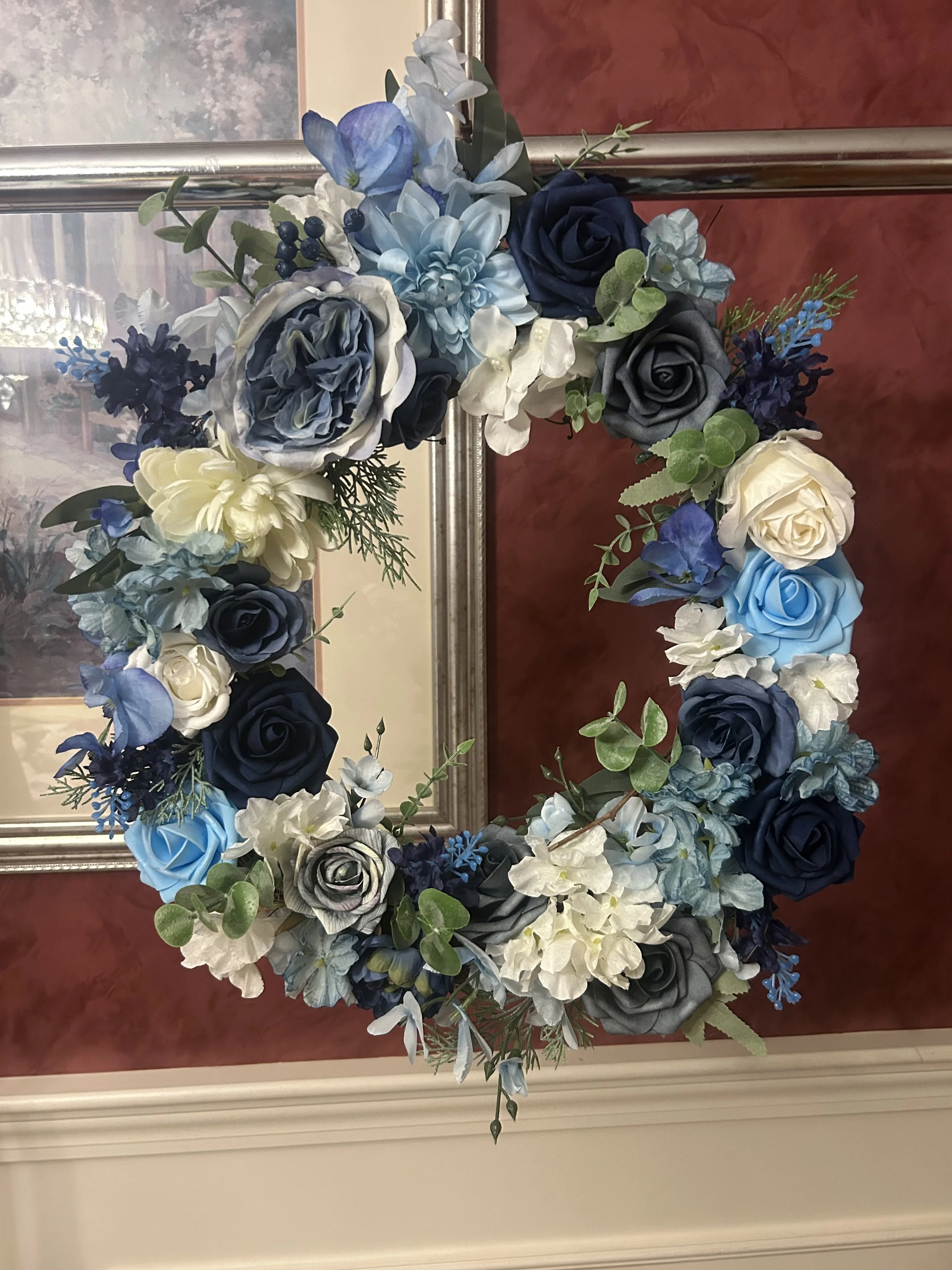A decorative floral wreath with blue, white, gray, and purple roses and various green foliage, hanging on a wall with a wooden frame mirror behind it.