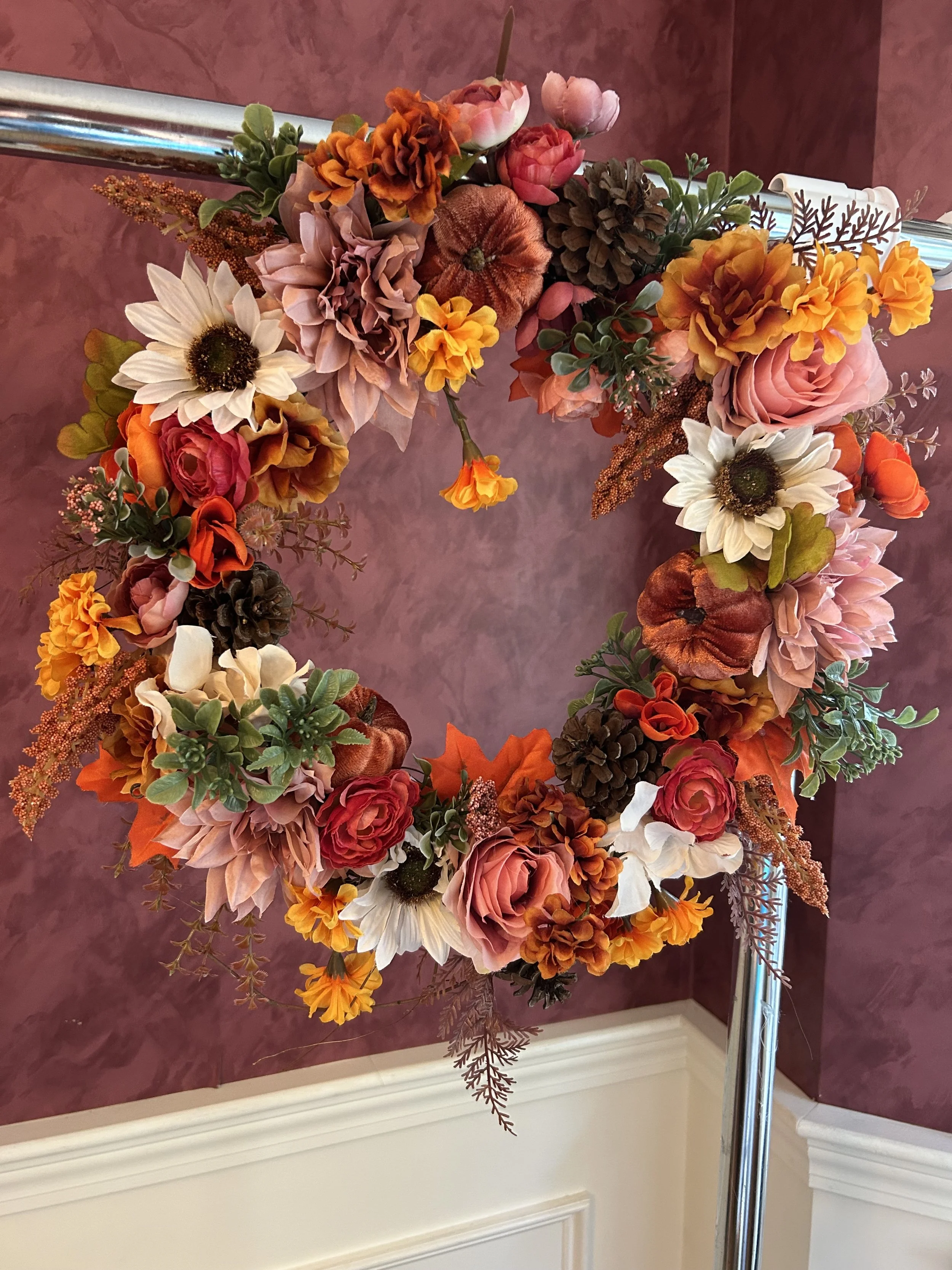 A floral wreath with various artificial flowers including sunflowers, roses, marigolds, and a pinecone, attached to a metal stand against a textured mauve wall.