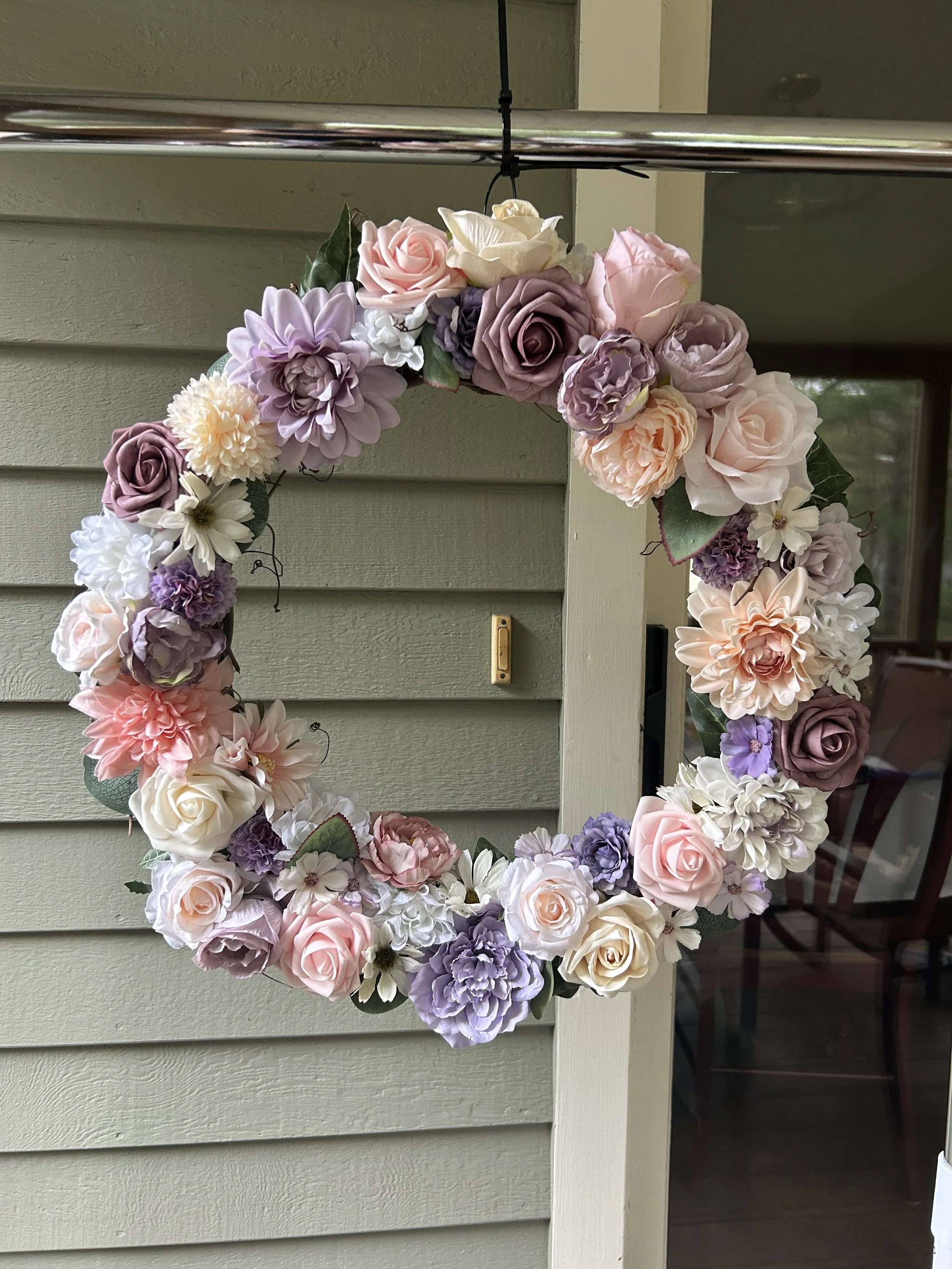 A floral wreath made of various pink, purple, white, and cream-colored artificial flowers hanging on a hook outside a house on a wall with horizontal siding.