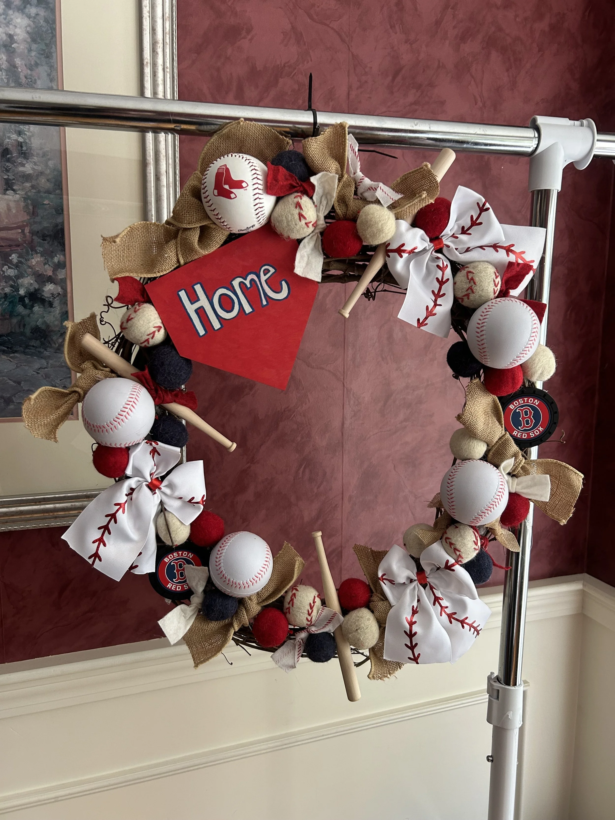 A decorative wreath featuring baseball-themed elements, felted baseballs, ribbons with baseball stitching patterns, burlap bows, and a small sign reading 'Home'.