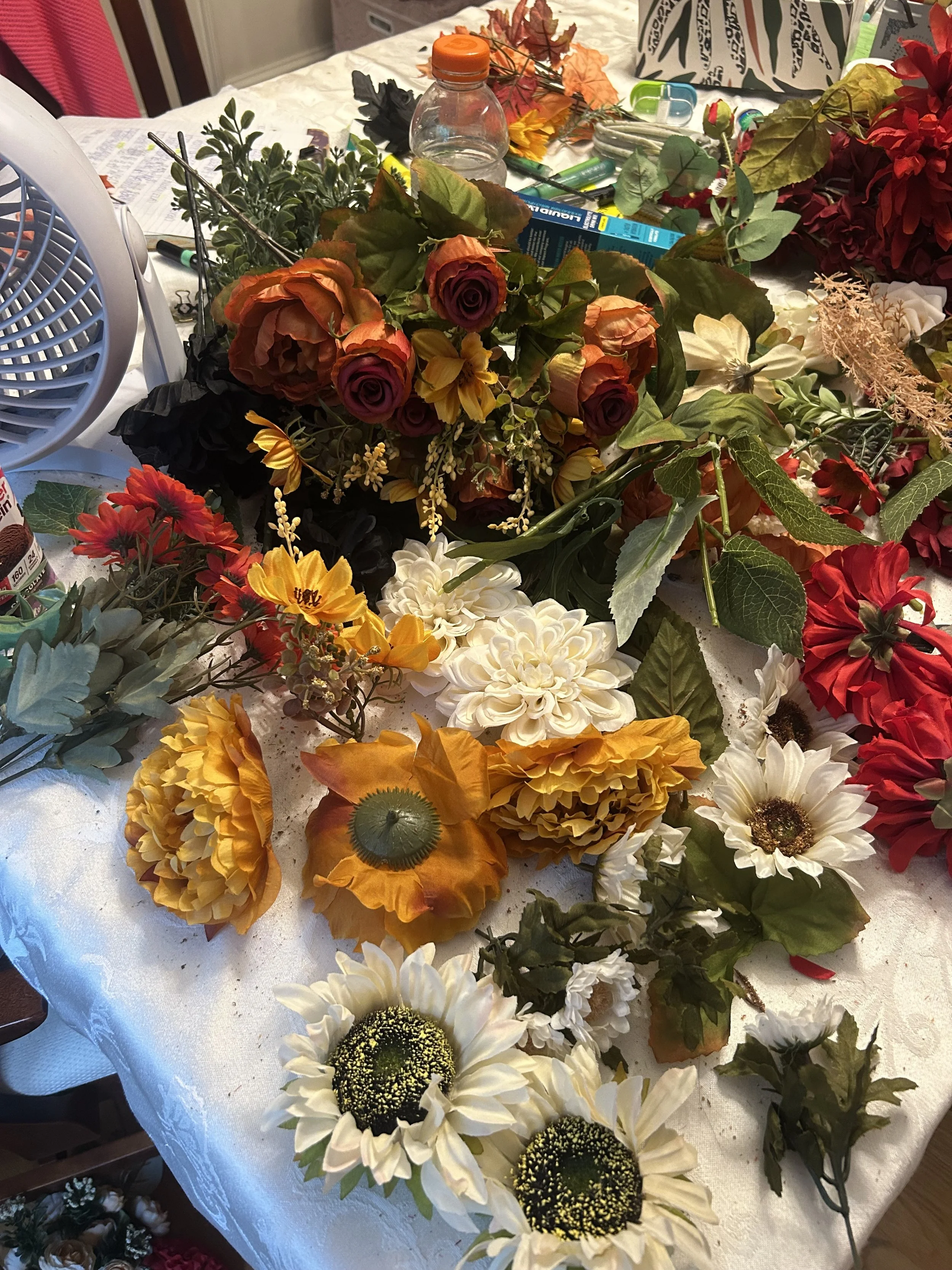 A cluttered table with artificial flowers, including white, yellow, orange, and purple roses and daisies, surrounded by various crafting supplies like glue, scissors, and packaging.