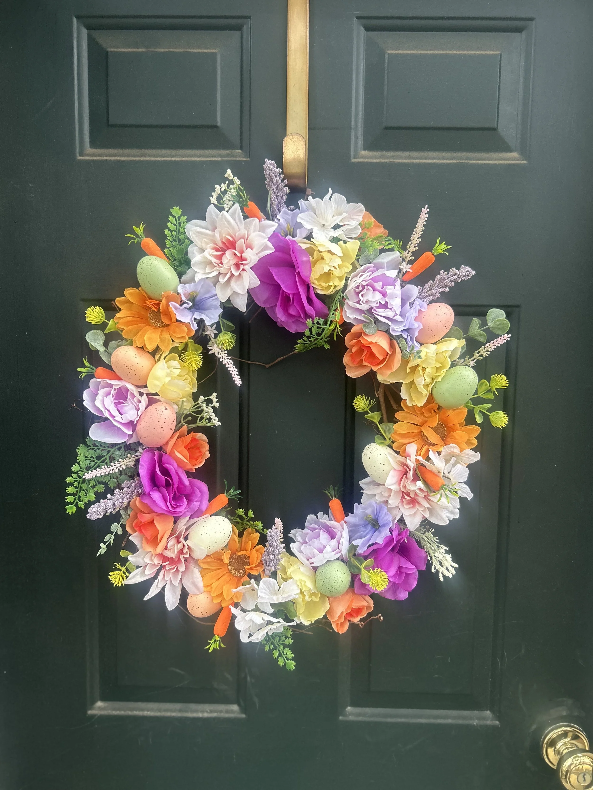 Decorative Easter wreath hanging on a dark green door, featuring colorful artificial flowers, pastel-colored speckled eggs, and small orange carrots.