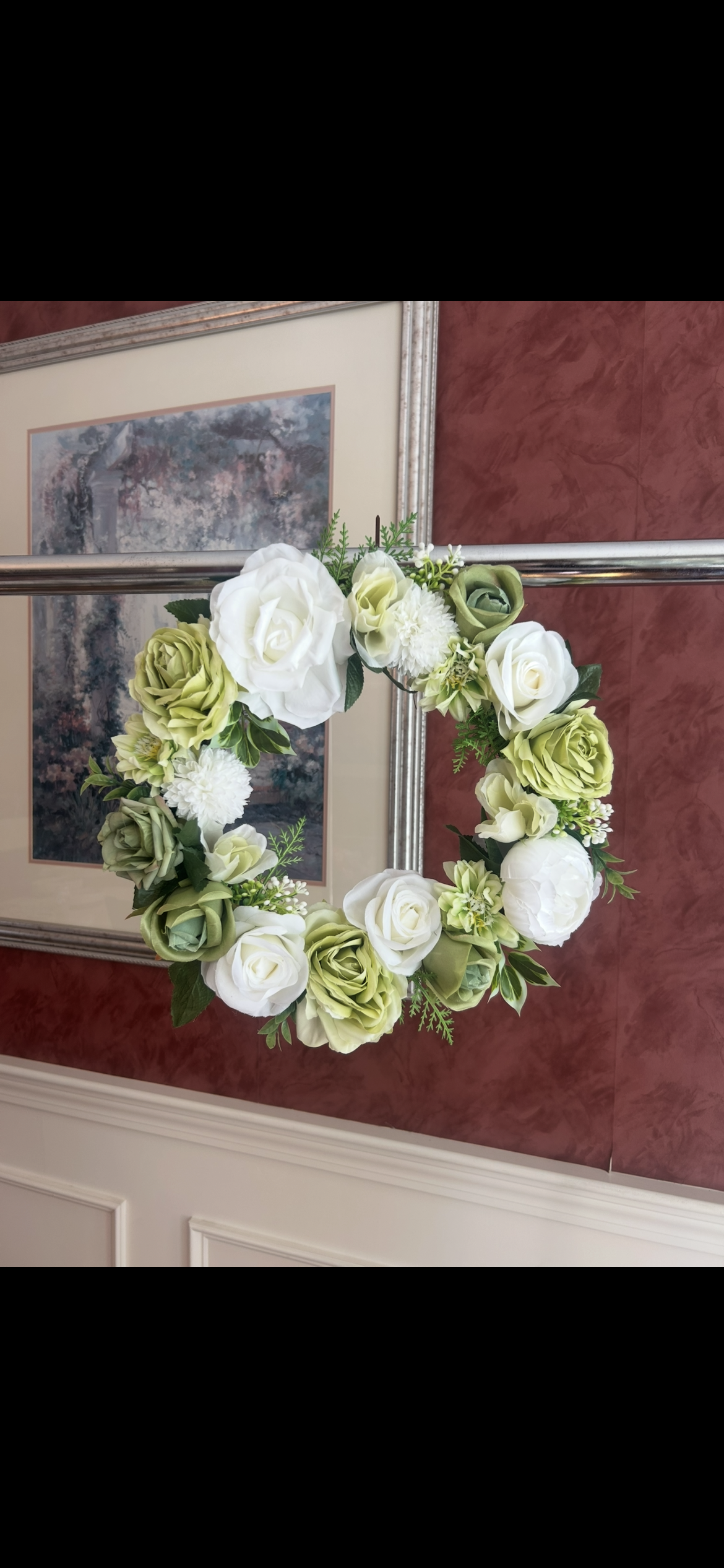 A floral wreath with white and green flowers mounted on a silver pole, with framed artwork and a red textured wall in the background.