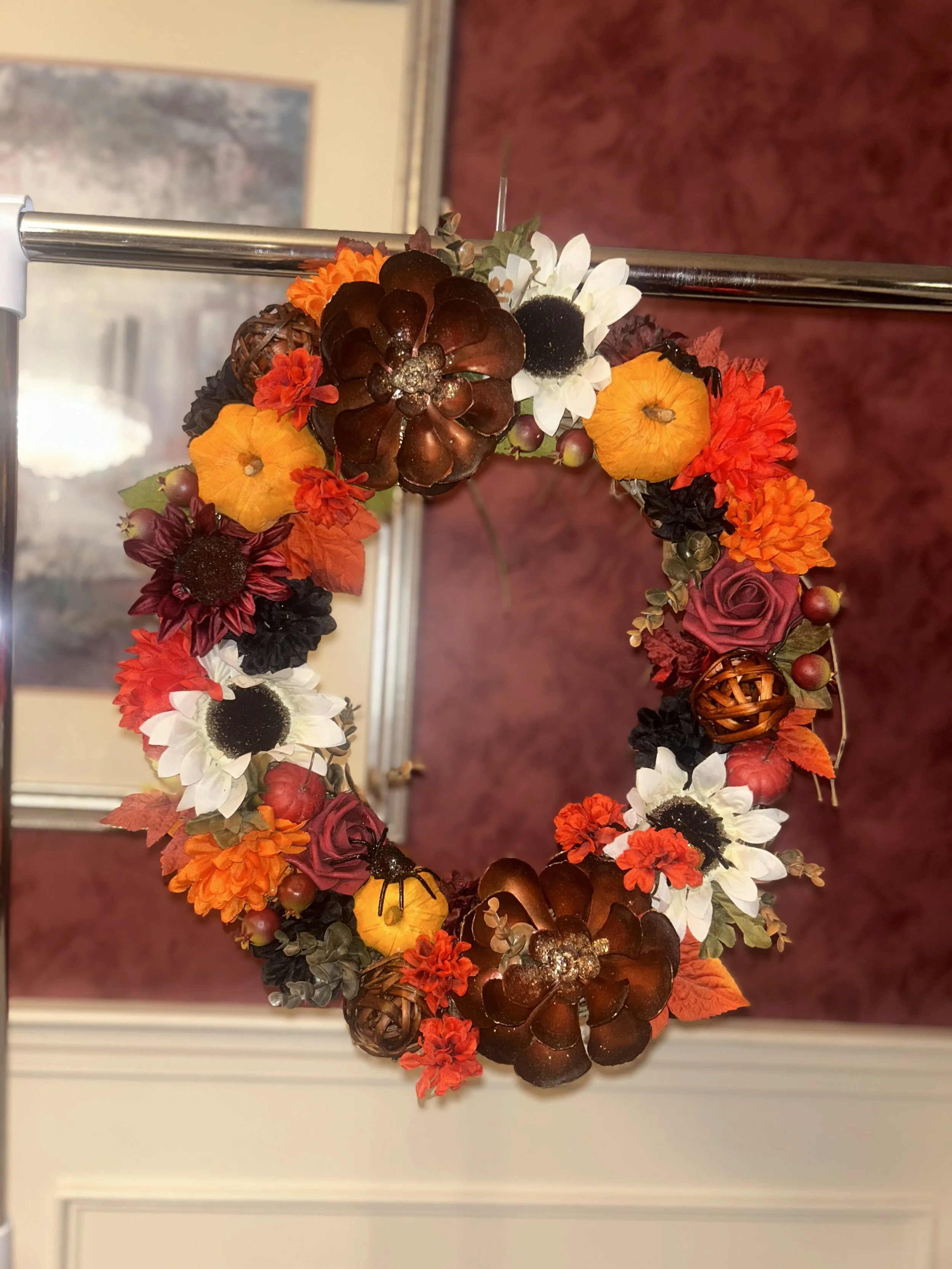 Decorated fall wreath with orange, white, yellow, red, and dark purple flowers, and various decorative elements, hanging on a metal pole.