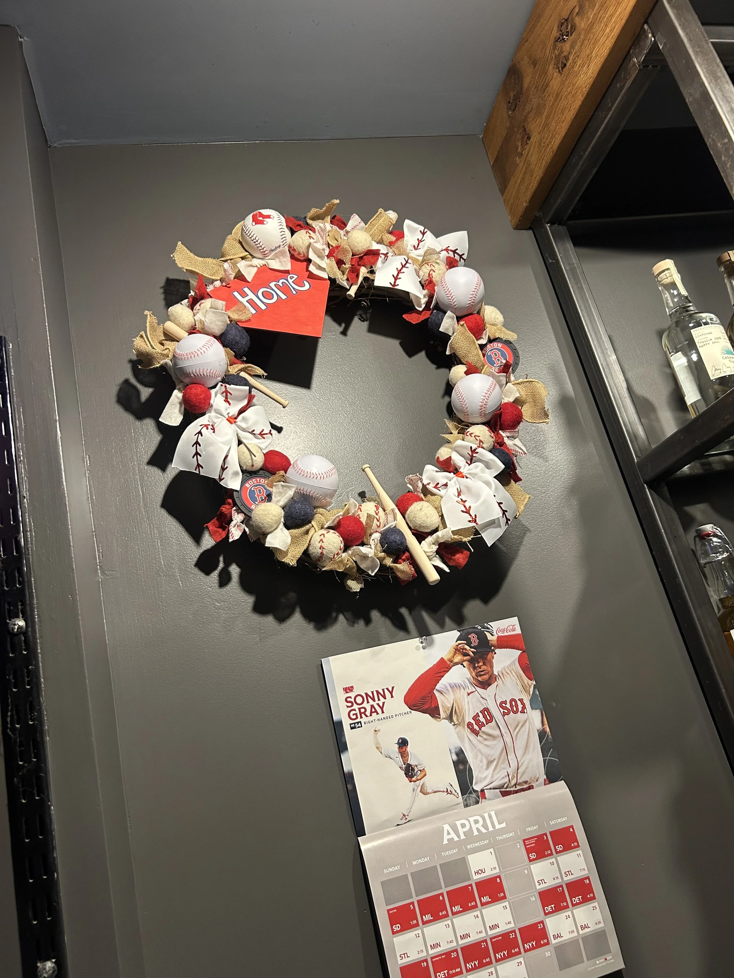 A decorative baseball-themed wall wreath hanging on a gray wall, featuring baseballs, fabric bows, and a red wooden sign with the word 'Home'; below is a calendar page for April with a photograph of a baseball player in a red uniform.