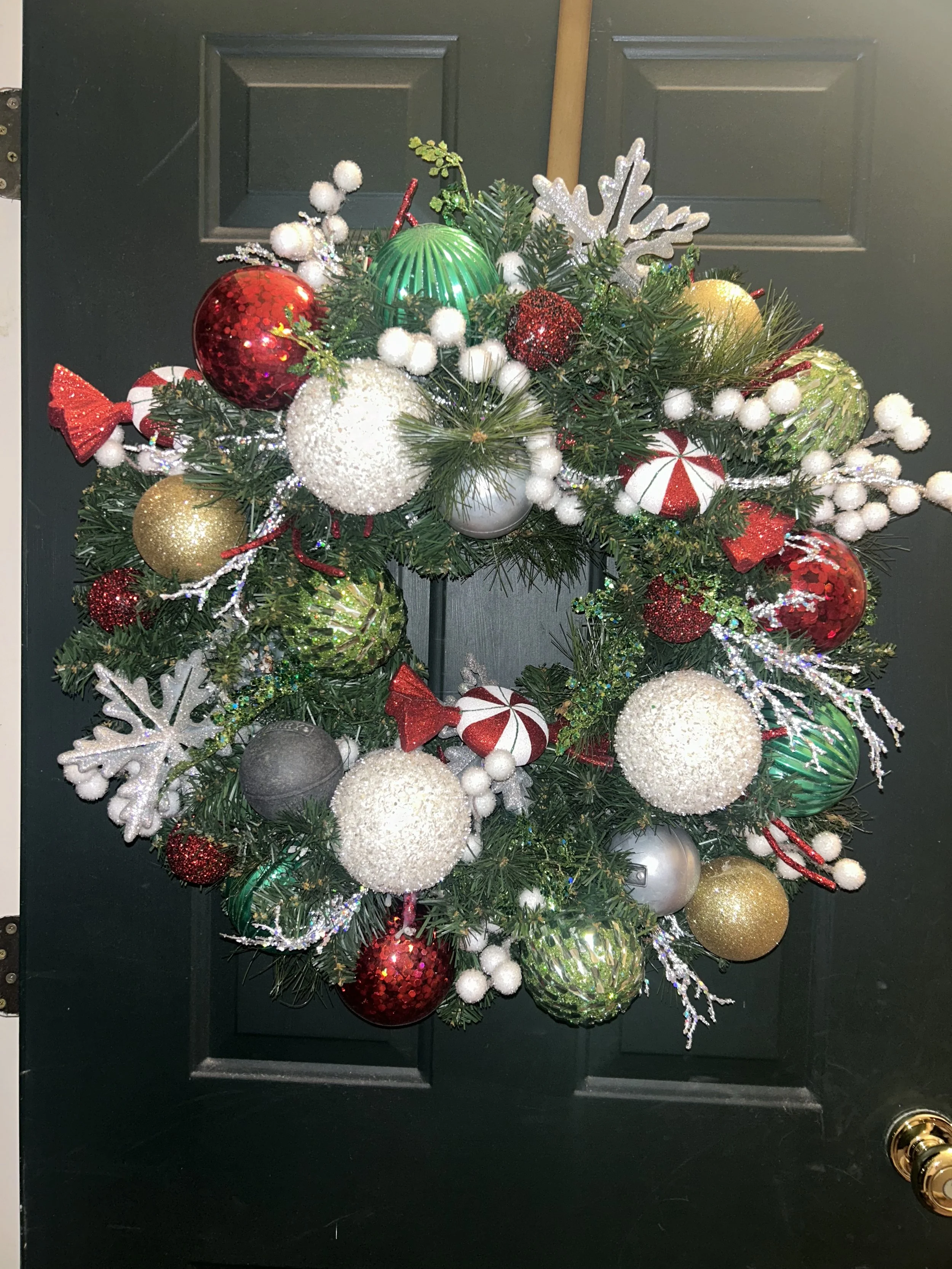 Holiday wreath decorated with red, gold, green, silver, and white ornaments, ribbons, snowflakes, and berry accents, hanging on a black door.