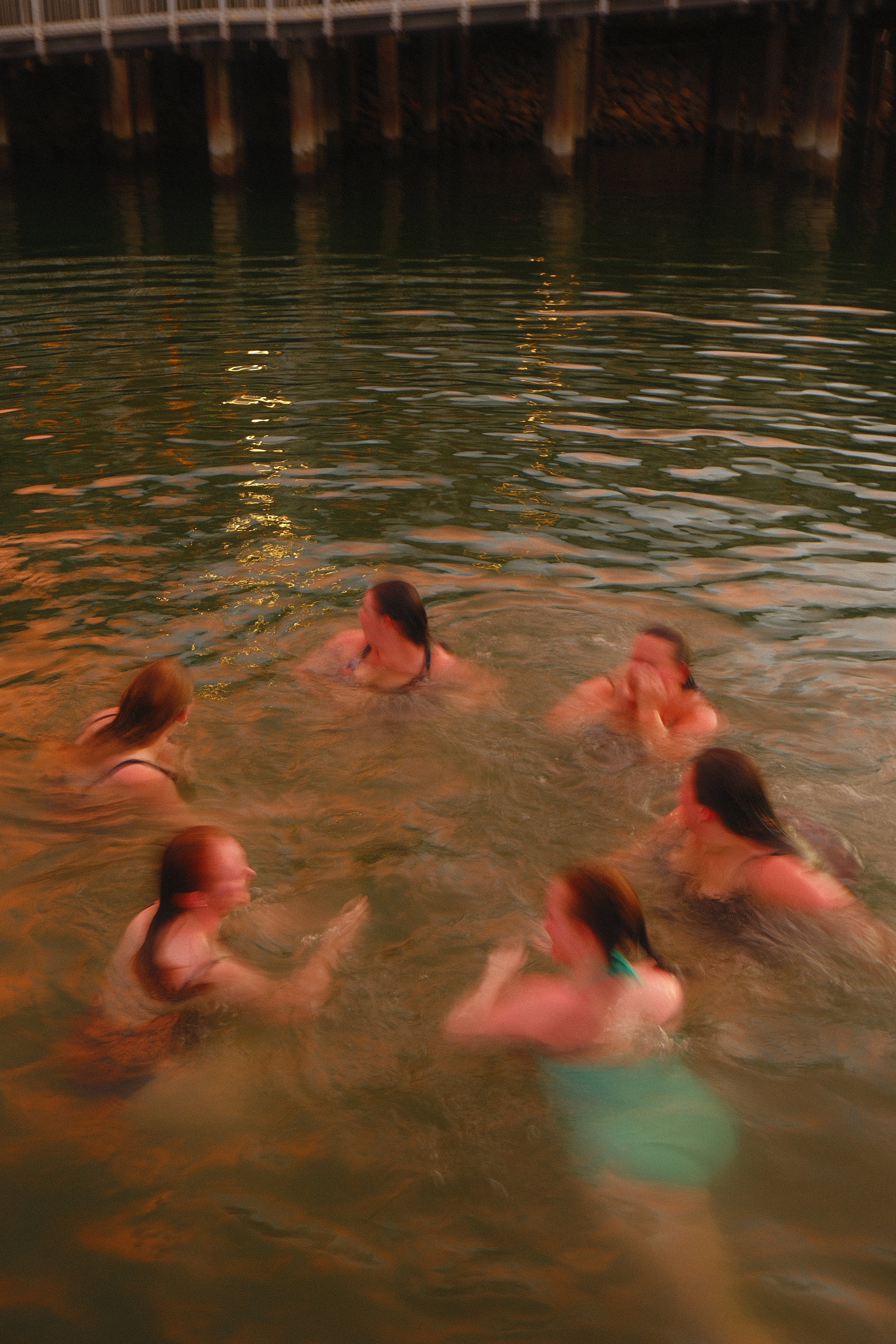 People swimming in a body of water near a bridge or pier at sunset or dusk.