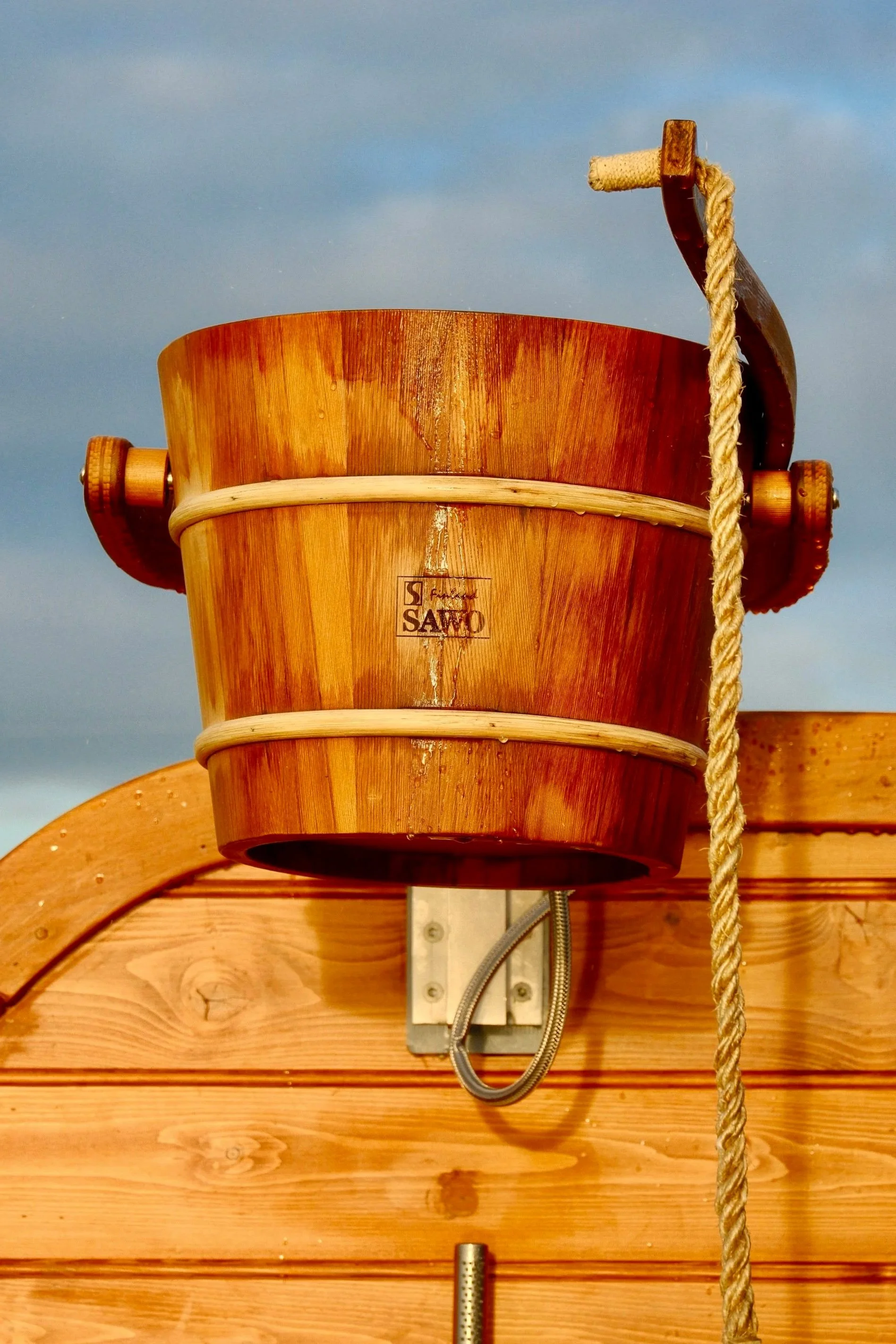 Close-up of a wooden bucket with a rope hanging beside it, mounted on a wooden wall with visible screws and a metal hook underneath, against a partly cloudy sky.