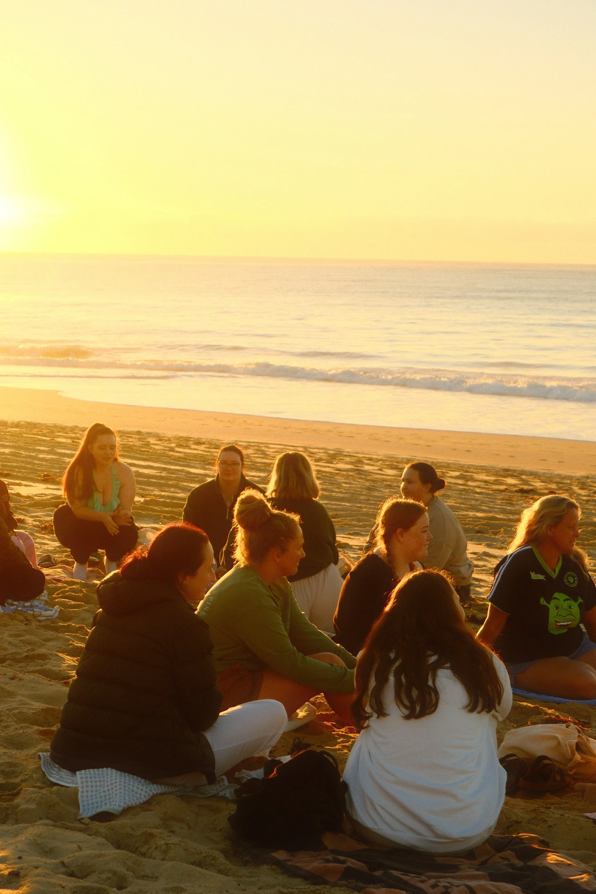 People sitting on a sandy beach during sunset, some in meditation or yoga poses, with the ocean and sky in the background.