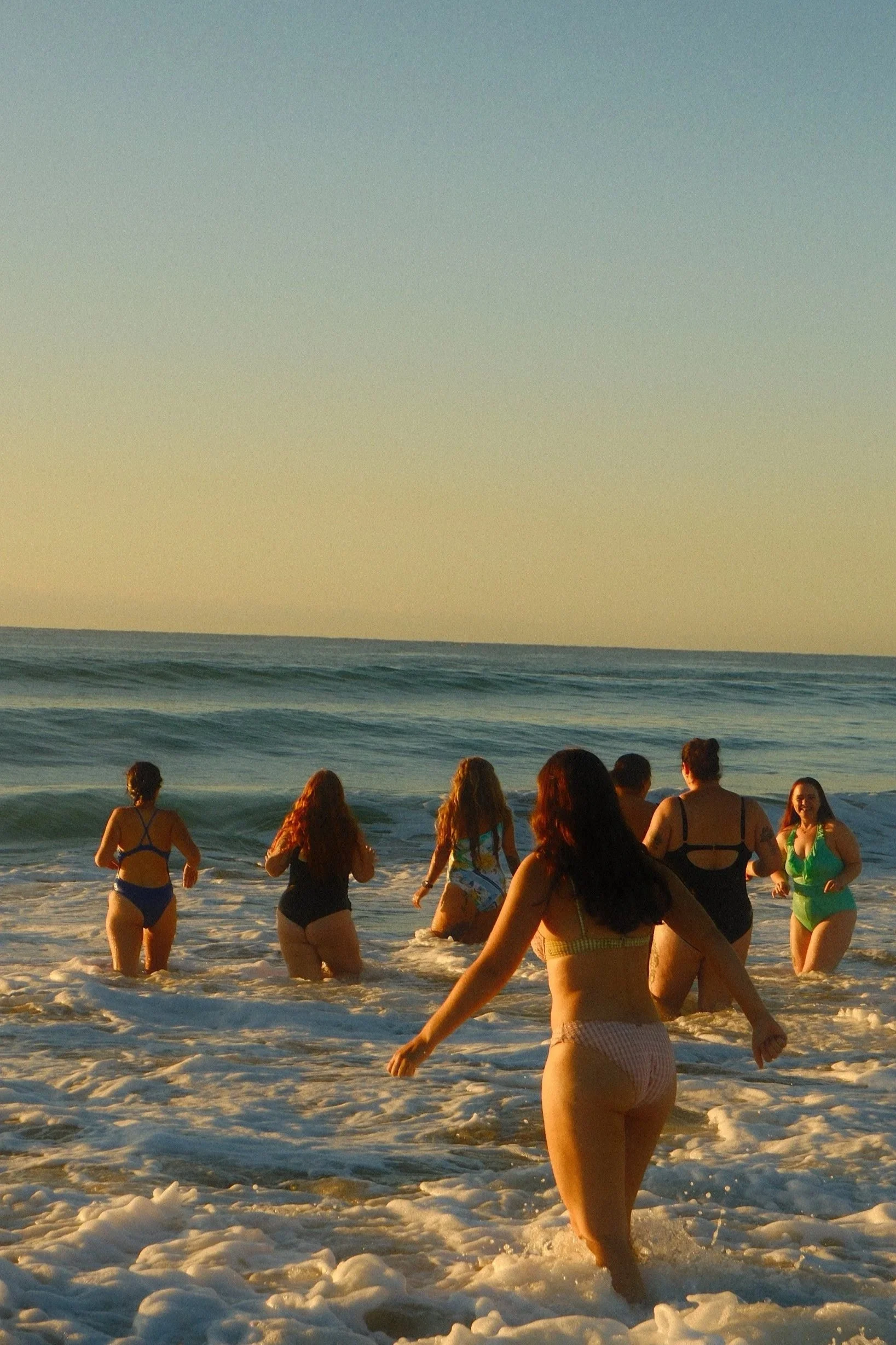 Group of people at the beach in swimsuits wading in the water, with the ocean and a sunset sky in the background.