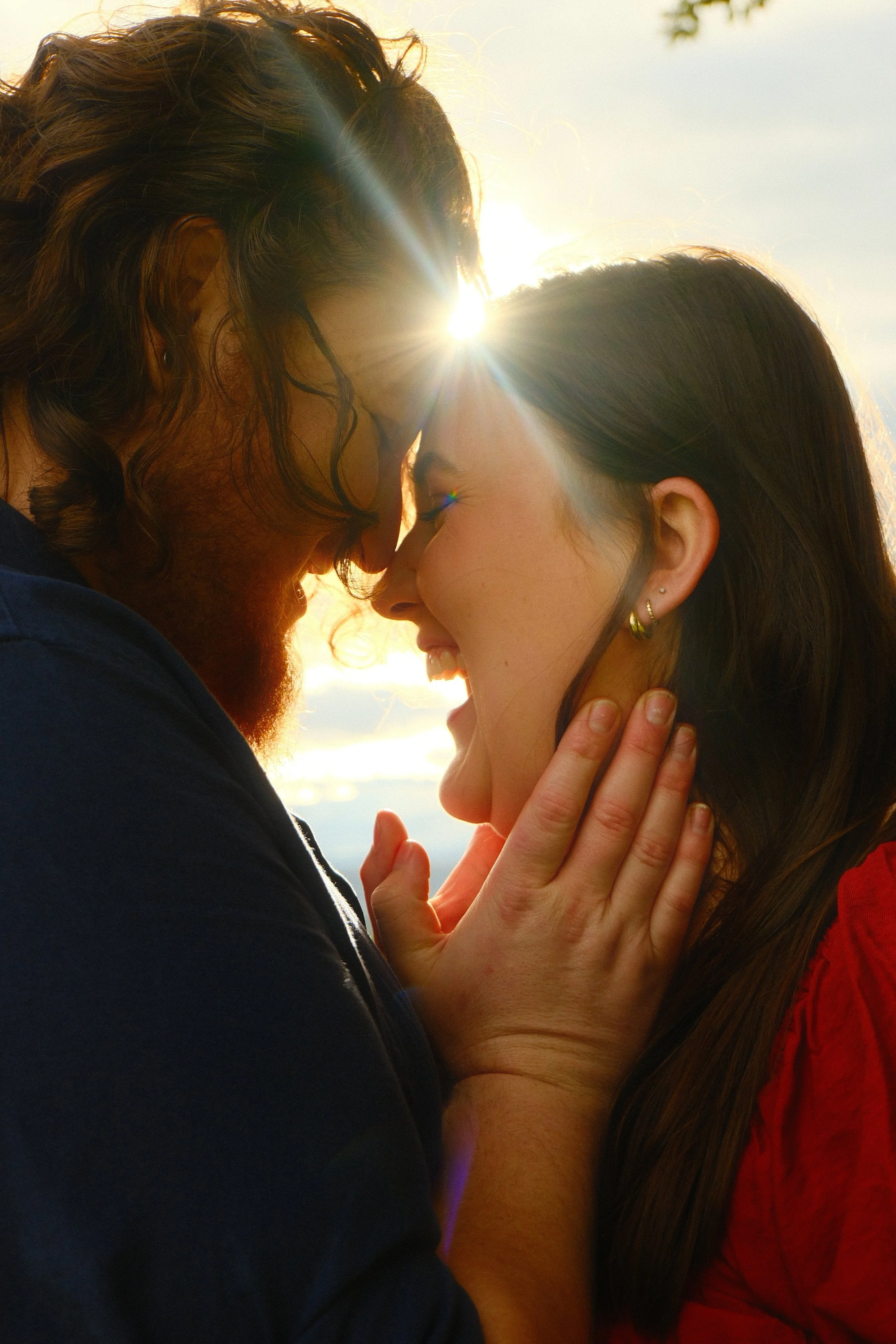 A couple with their foreheads touching, illuminated by the sun at sunset or sunrise, with the woman gently holding the man's face and both appearing happy and intimate.