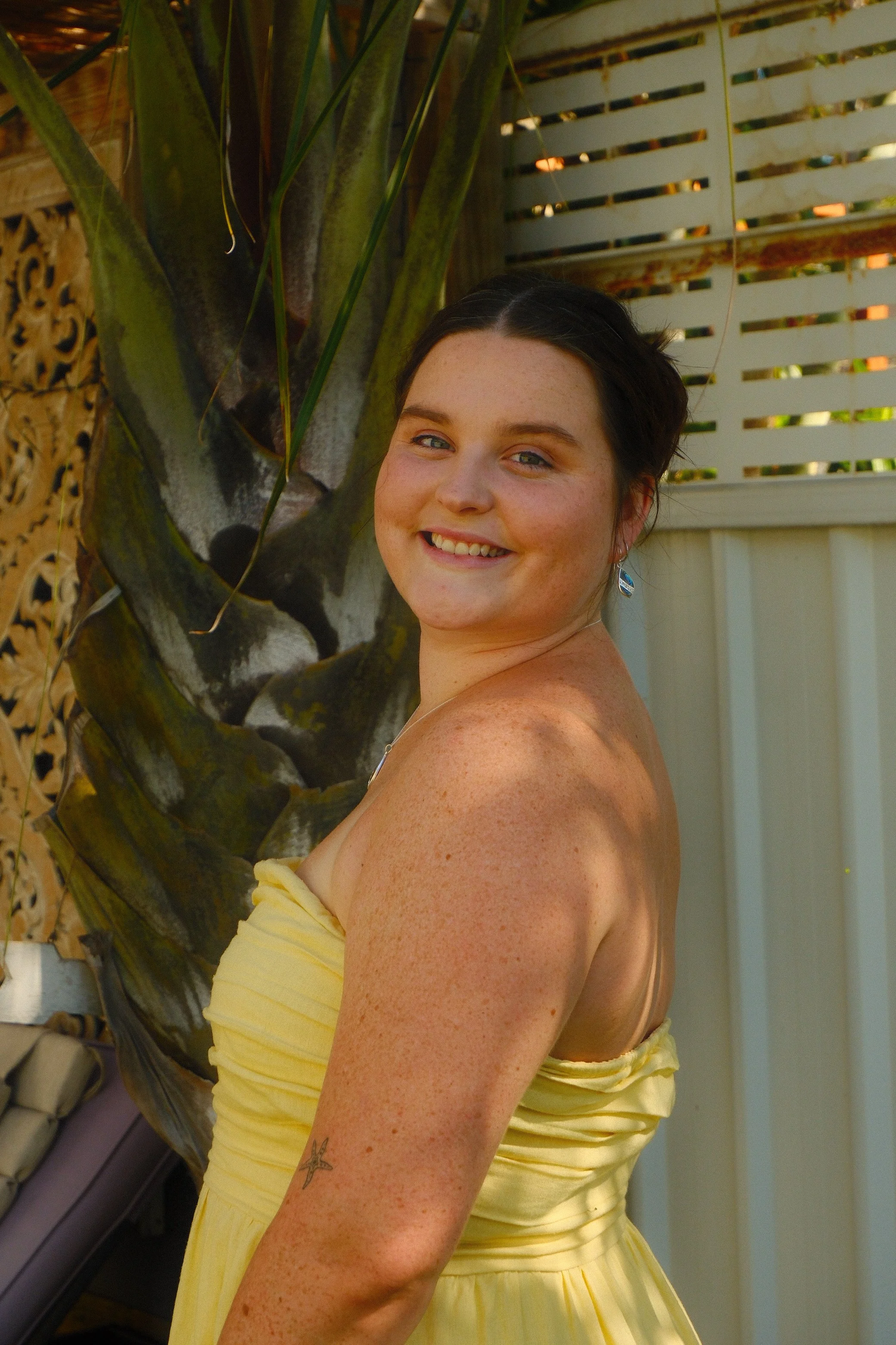 A young woman with dark hair, blue eyes, freckles, and a star tattoo on her arm, smiling and wearing a light yellow dress, standing outdoors near a palm tree and a wooden fence.