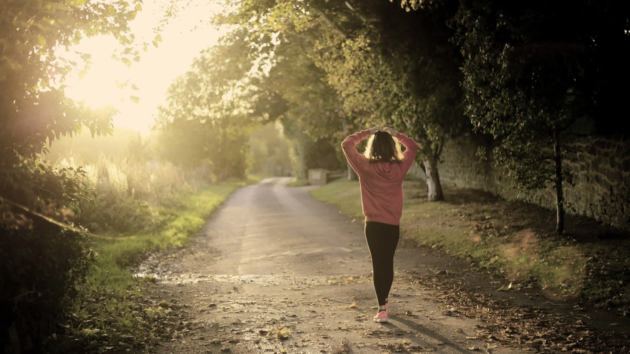 A person, wearing a pink hoodie and black leggings, is walking on a dirt path surrounded by trees, with the sun shining through the leaves, creating a warm, glowing atmosphere.