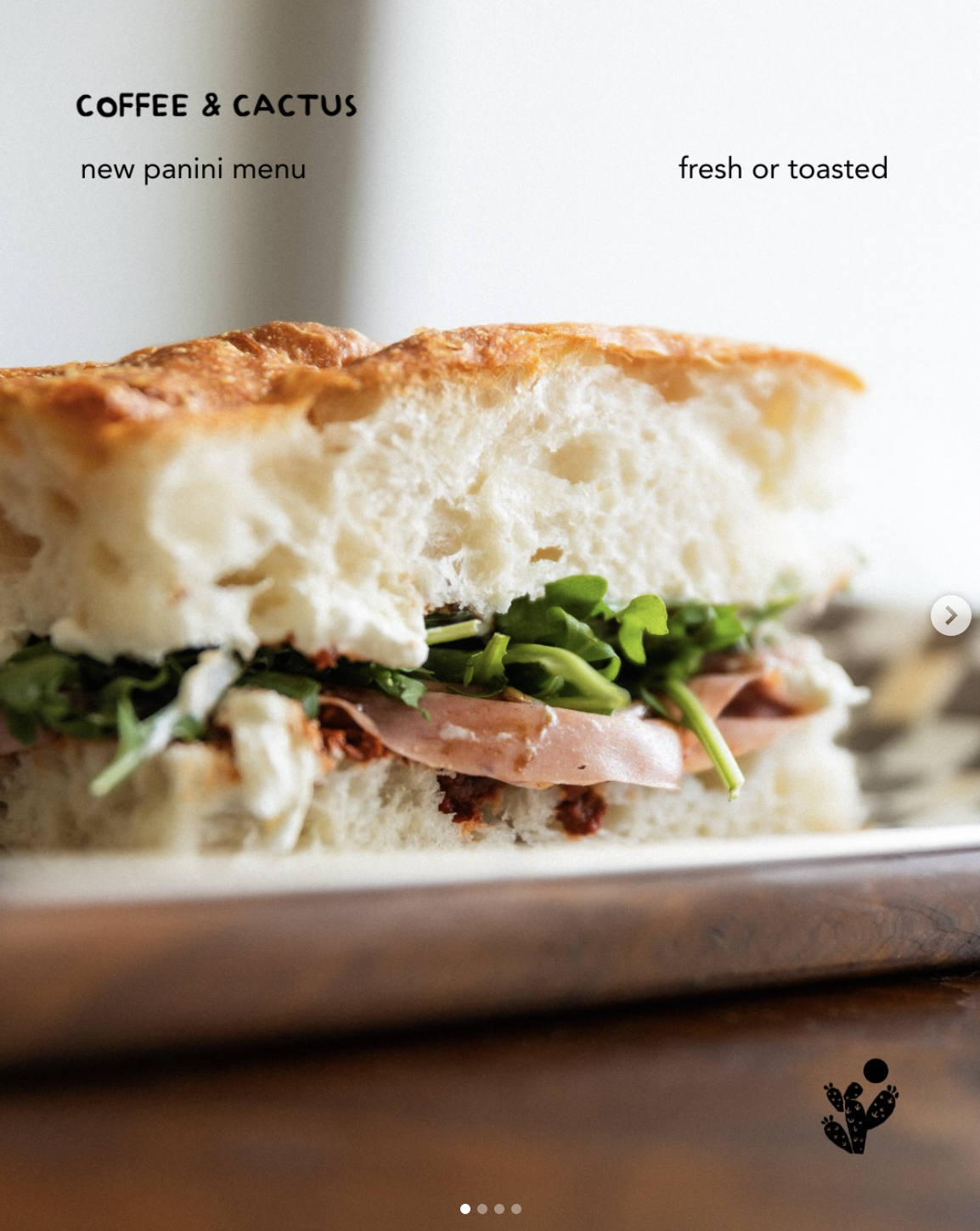 Close-up of a sandwich with bread, lettuce, and ham, placed on a wooden surface. The background features a white wall with text about a new panini menu from Coffee & Cactus.