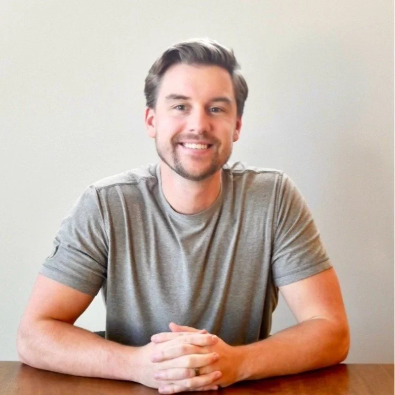 Smiling man with brown hair and beard, wearing a gray t-shirt, sitting at a wooden table in front of a plain light-colored wall.