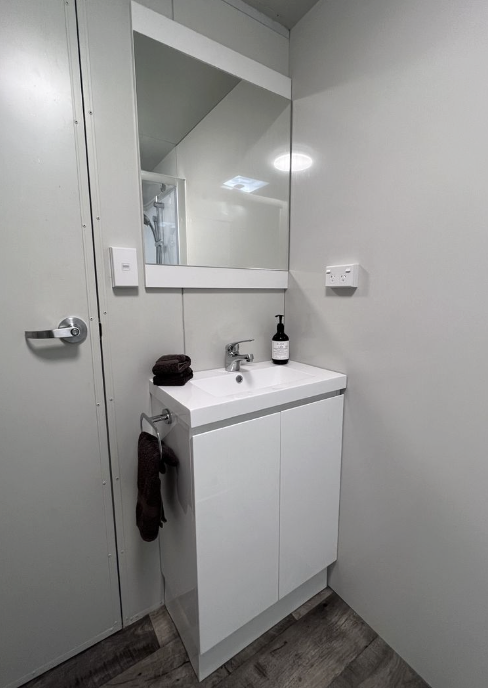 Small white bathroom sink with a black soap dispenser on top, a dark brown towel on the sink, and a dark hand towel hanging from the side of the cabinet, with a large mirror above it