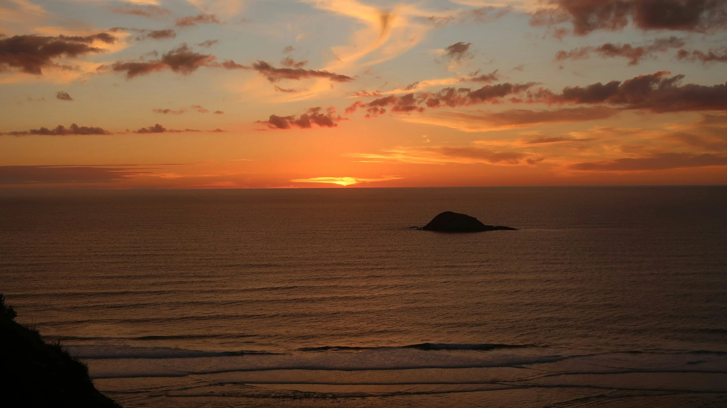 A scenic ocean view at sunset with an orange and pink sky, some clouds, calm water, and a small island or rock formation in the distance.