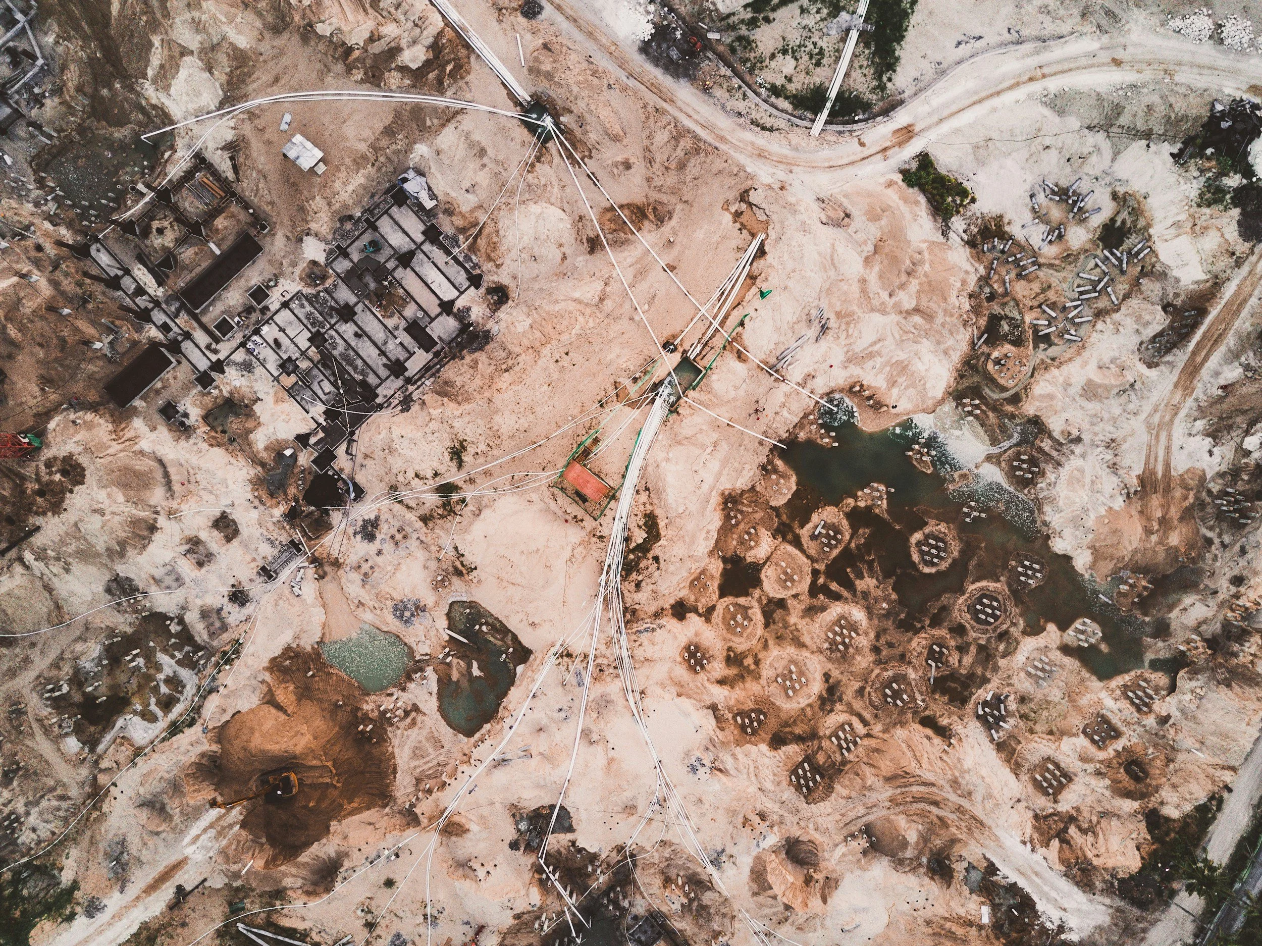 An aerial view of a construction site with exposed earth, construction materials, and equipment. Several pipes and cables are laid out across the site, and there are partially built structures and water bodies within the excavation area.
