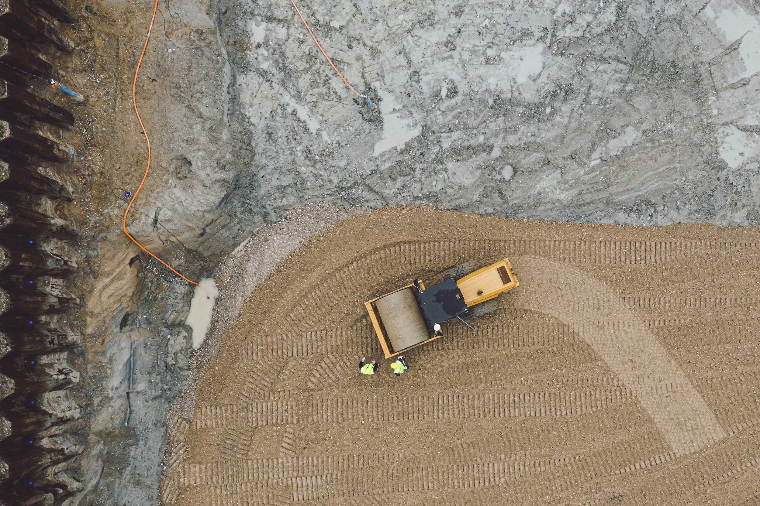 Aeroshot of a construction site showing an excavated area with three workers in high visibility jackets and helmets, a small orange dump truck, a construction roller, and orange safety cables, with a gravel and dirt surface.