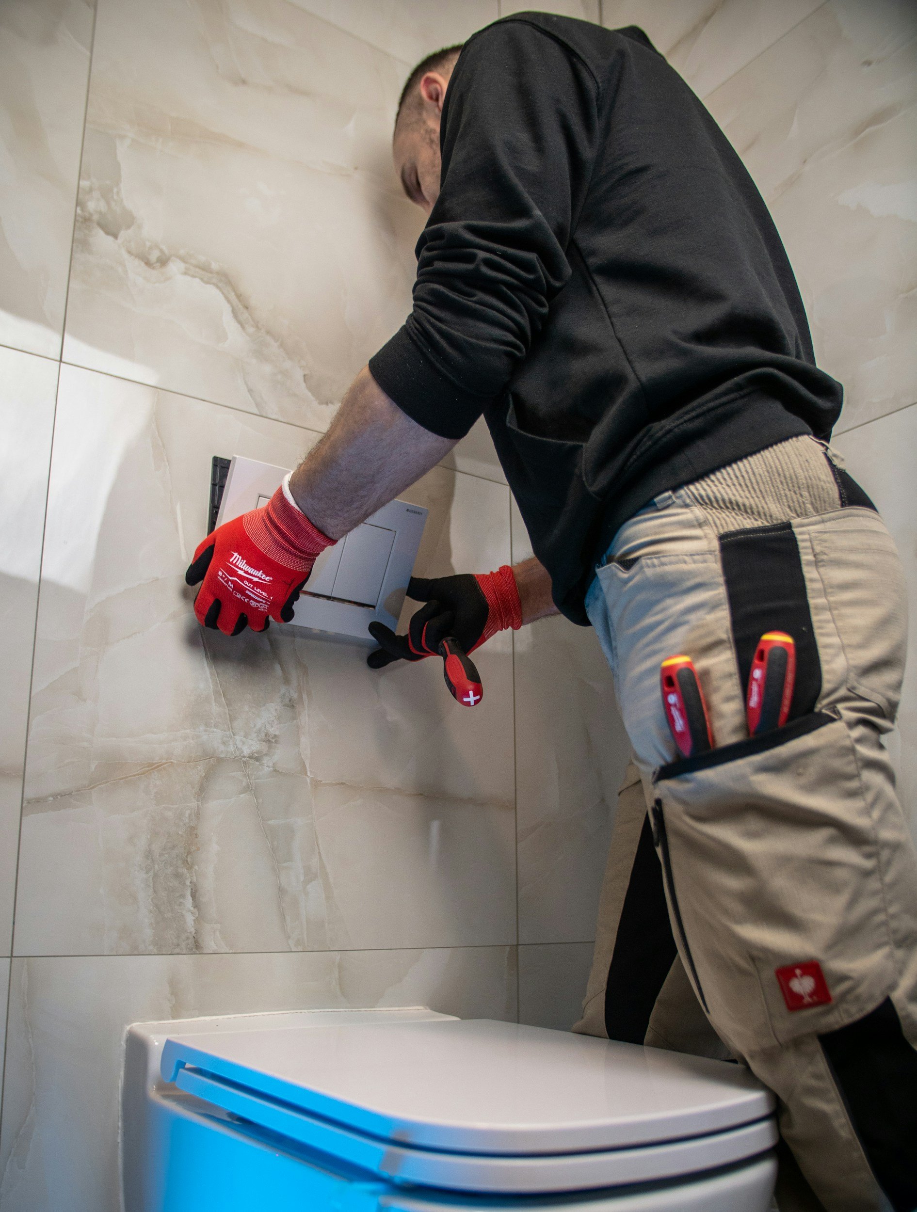 A man installing or repairing an electrical outlet on a beige marble wall, wearing red Milwaukee work gloves and beige work pants with a Swiss flag patch, in a bathroom or kitchen.