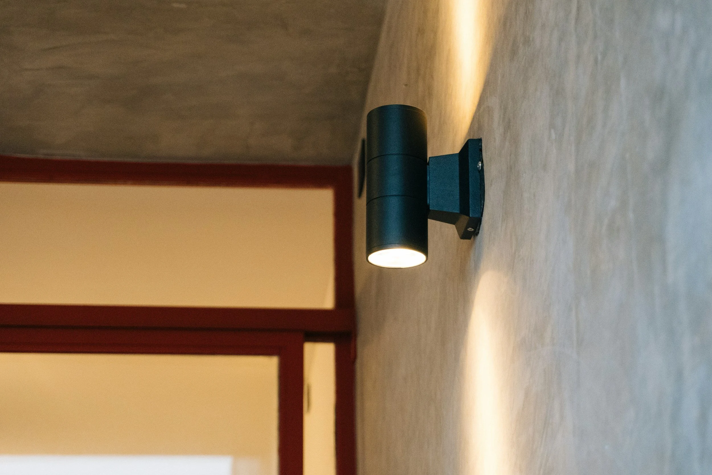 A modern black wall-mounted cylindrical light fixture illuminating a wooden surface on a wall. The floor appears to be a textured material, and there is a vertical red-brown wooden frame on the left side of the image.