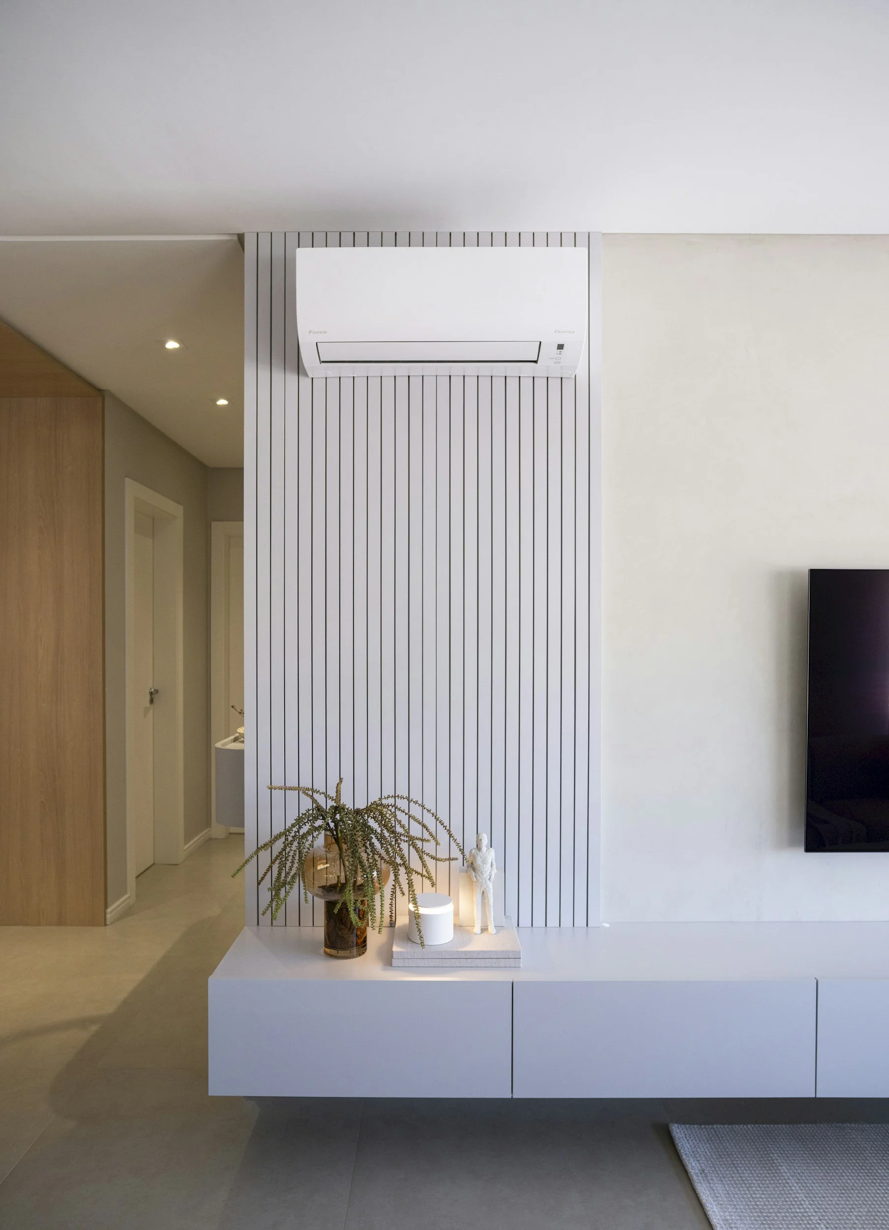 Modern interior with a white wall and a decorative setup on a white cabinet, including a large brown vase with greenery, a white sculpture, and a lit candle, against a striped wall panel in a minimalist space.