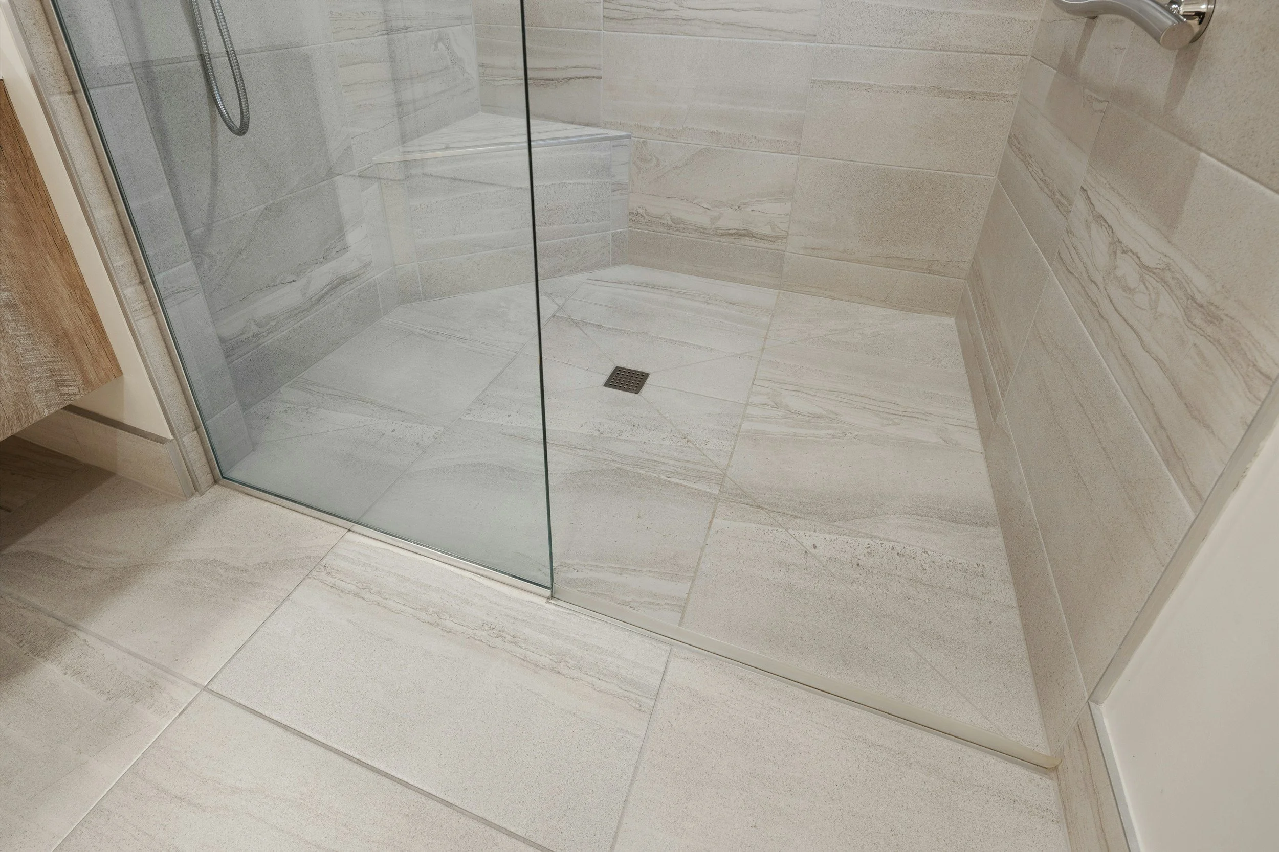 A corner of a bathroom shower with beige marble tiles, a glass shower door, a silver showerhead, and a floor drain.