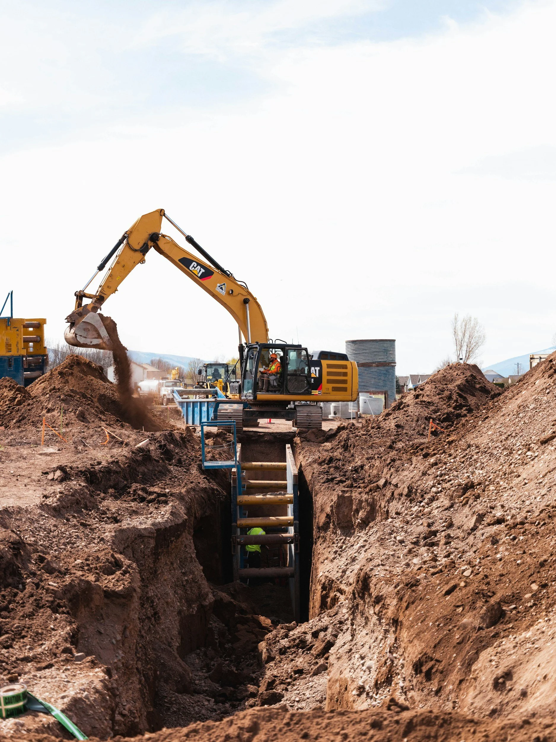 Construction site with excavator digging a trench, worker inside trench, and blue support beams.