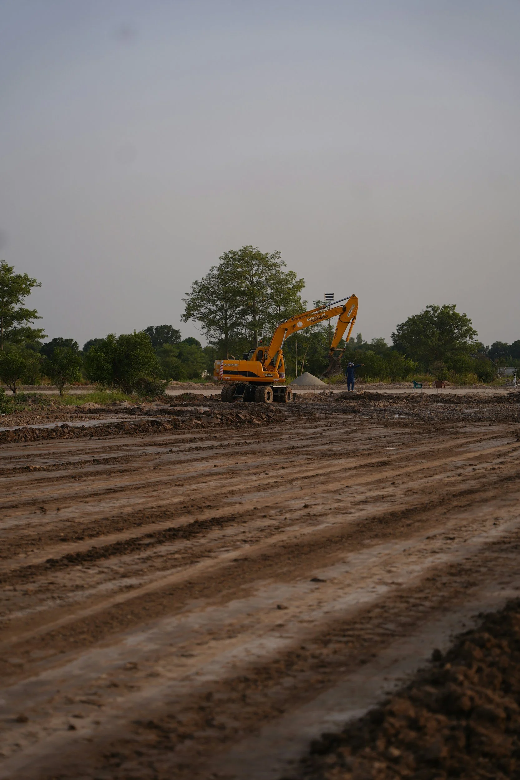 Construction site with excavator digging soil, trees in background under cloudy sky.