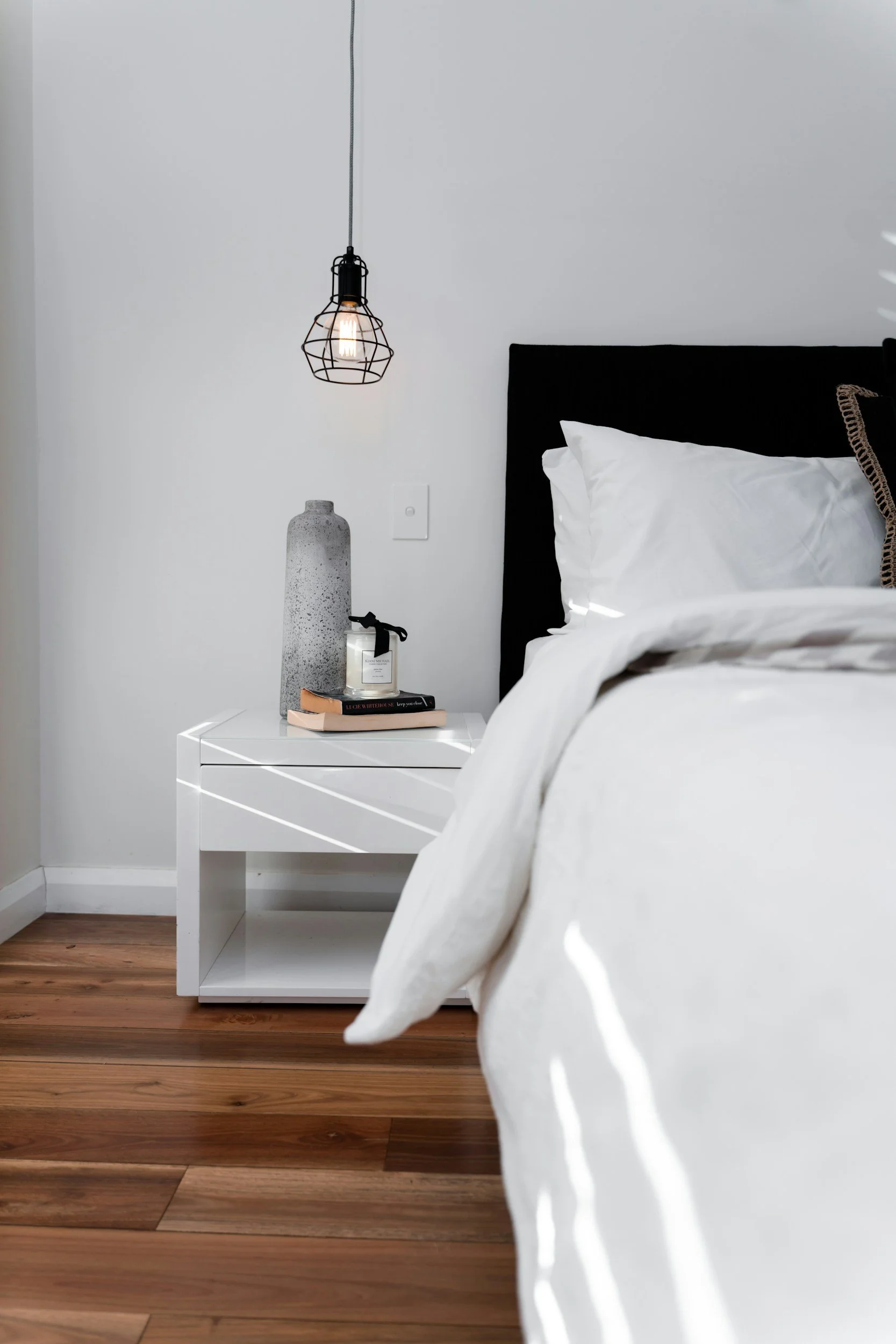 A bedroom corner featuring a white nightstand with books, a candle, and a tall textured vase, next to a bed with white bedding and a black headboard. There is a wall-mounted light fixture with a wire cage design hanging above.