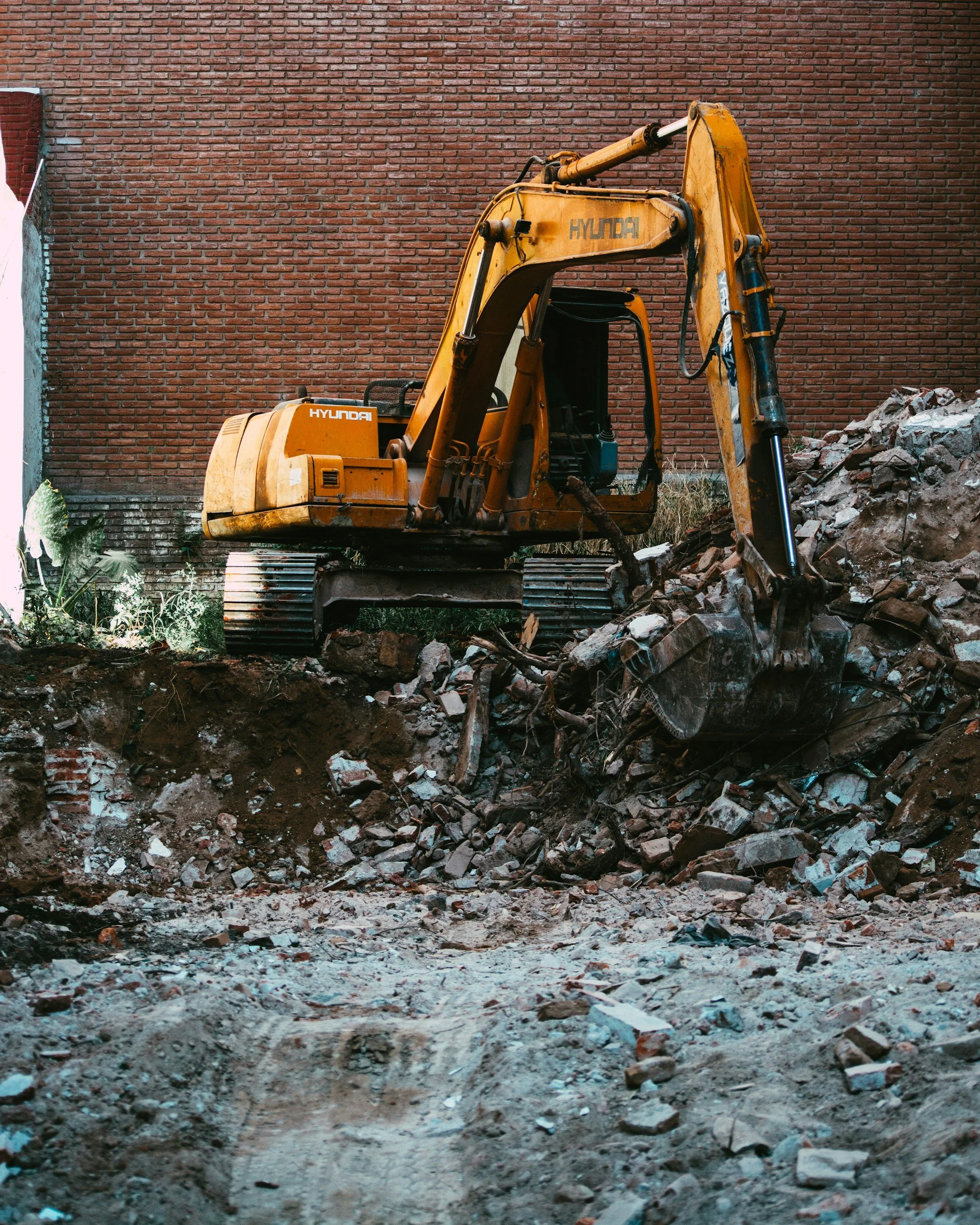 A yellow Hyundai excavator working on a construction site with a brick wall in the background and rubble in the foreground.