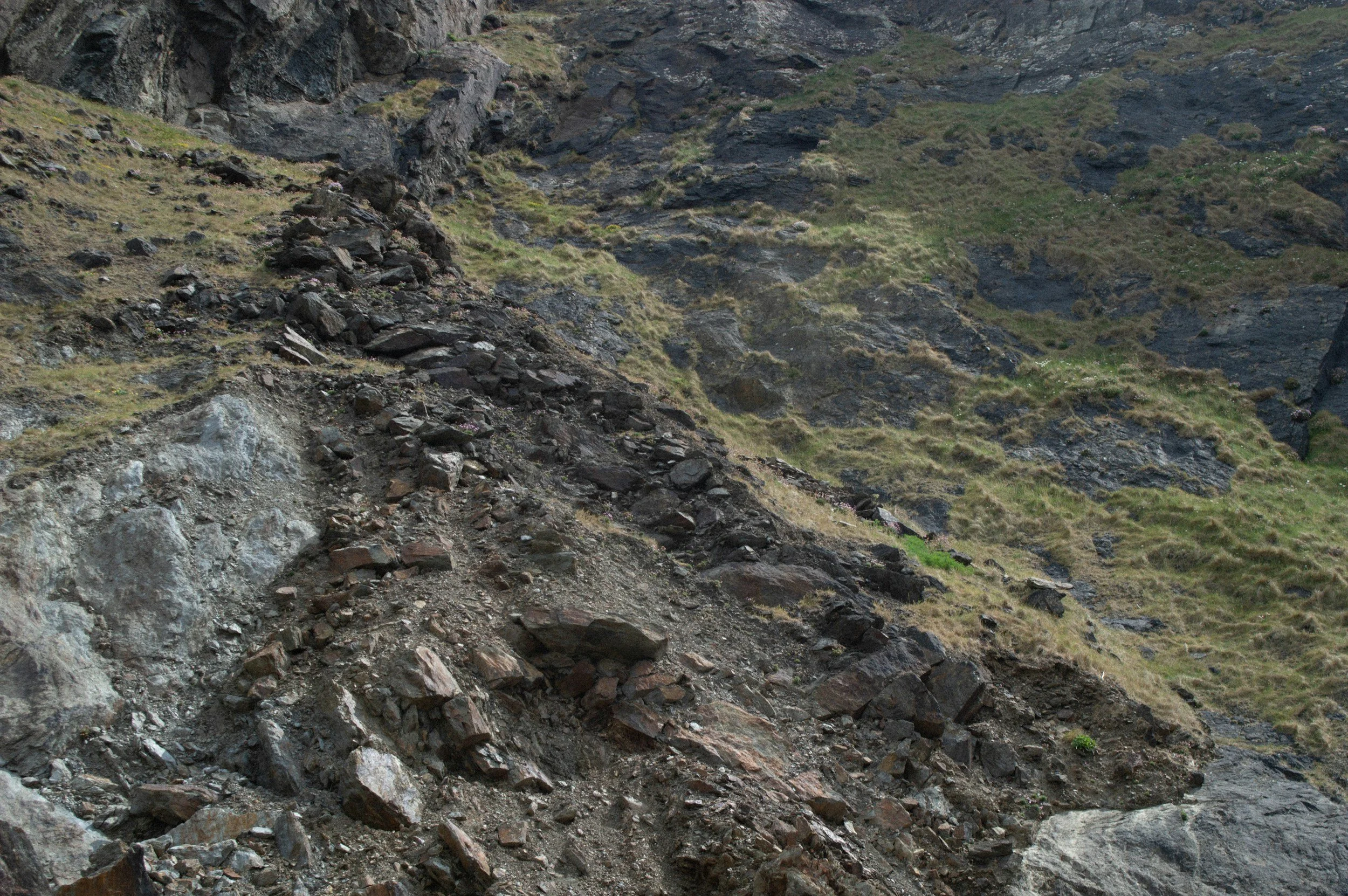 A rugged hillside with loose rocks and sparse patches of grass, showing a steep incline with rocky terrain.
