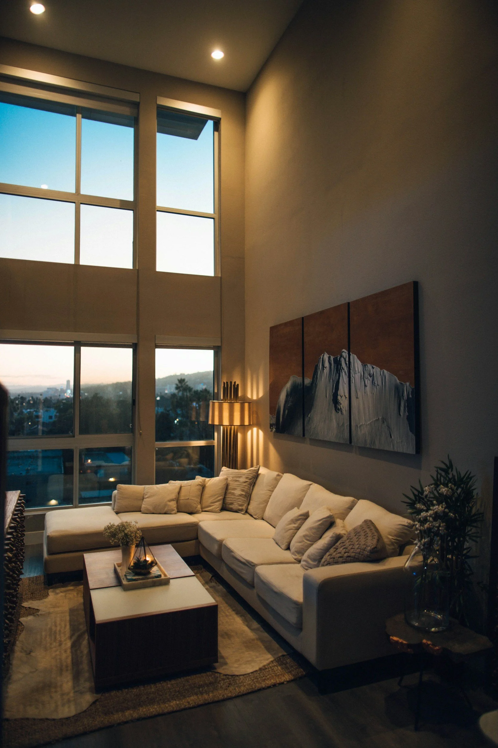 Living room with large windows showing a city view, beige sectional sofa with throw pillows, wooden coffee table with decorative tray, tall floor lamp, artwork on the wall, plant in vase, and cozy lighting.