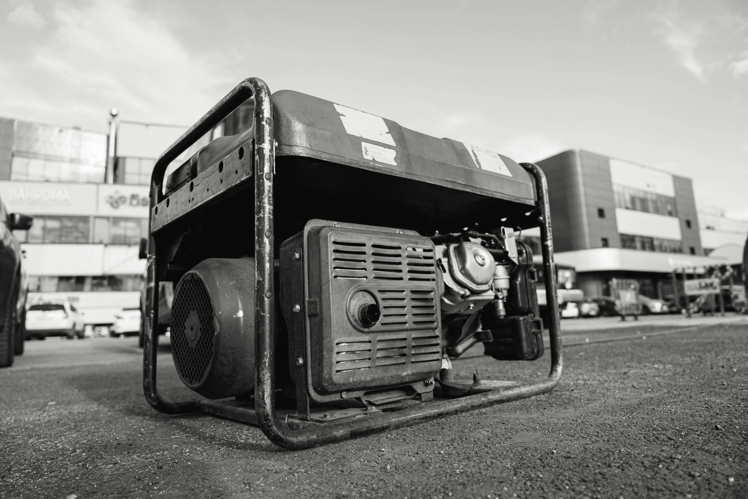 A black and white photo of a portable generator on a parking lot with a modern building in the background.