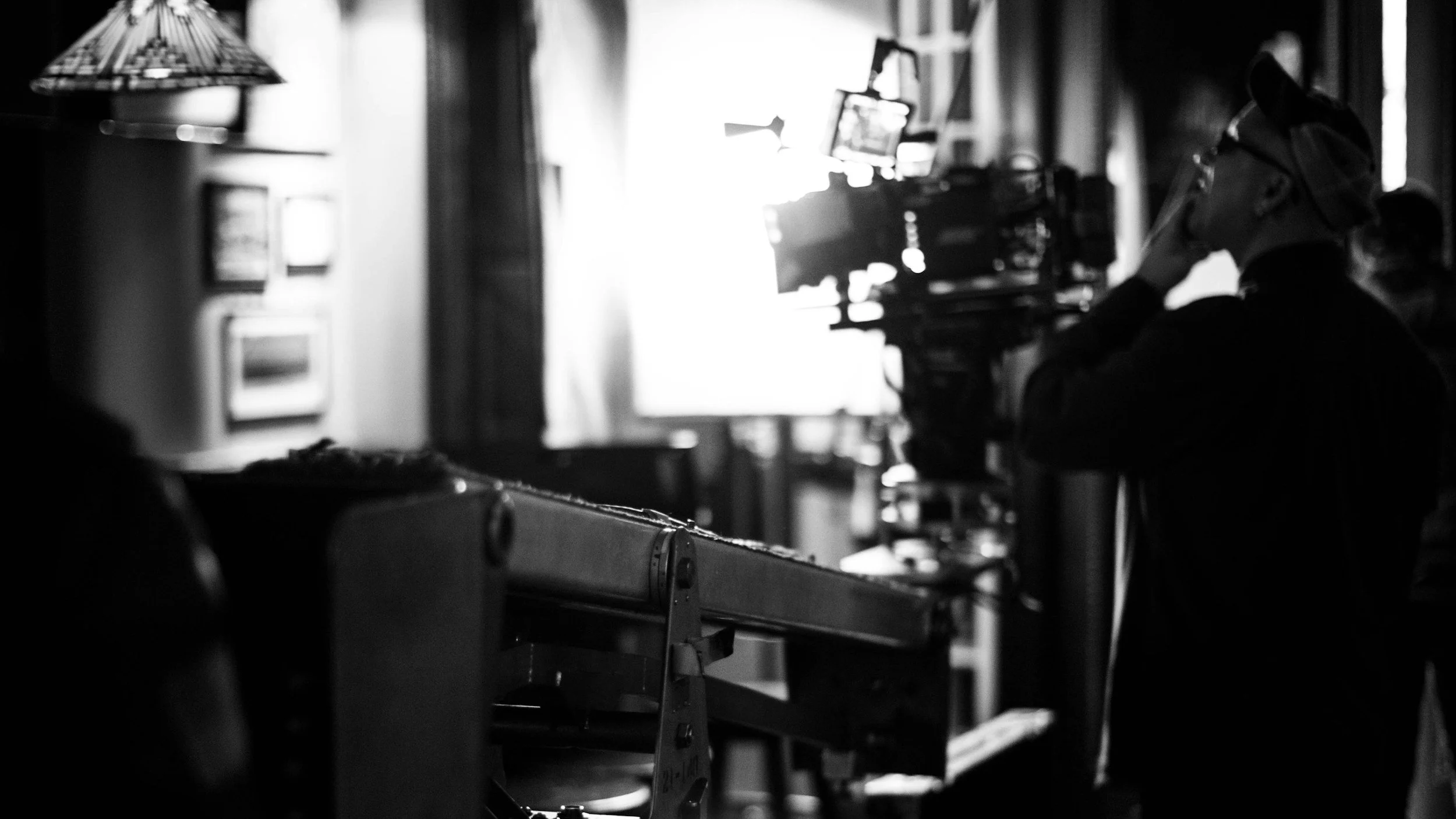 Black and white photo of a person working with a camera or filming equipment, seated in a room with curtains and framed pictures on the wall, bright light streaming in from a window.