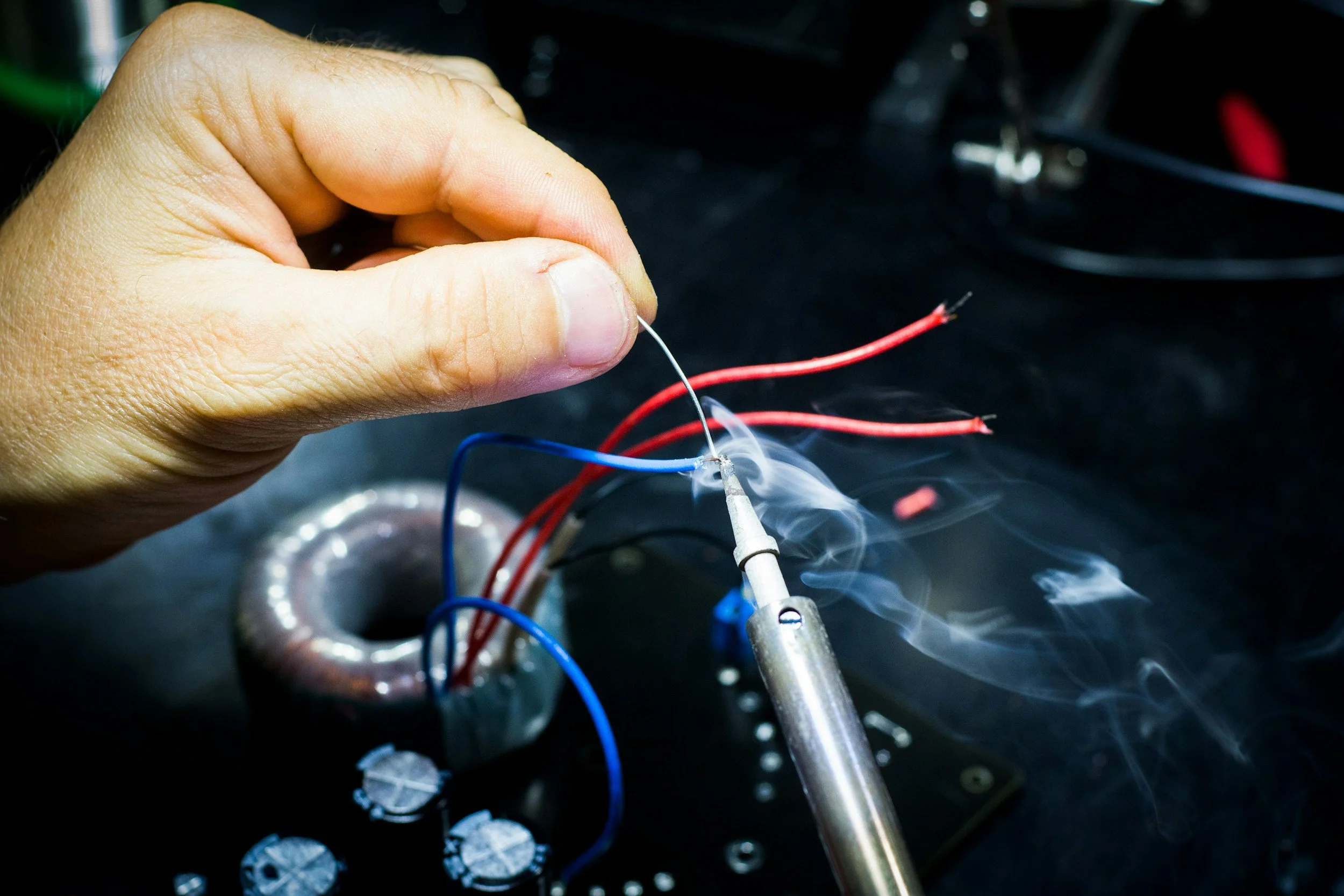 A person's hand holding a thin wire soldering it to a small electronic component with red and blue wires connected, on a dark surface with metallic and electronic parts scattered.