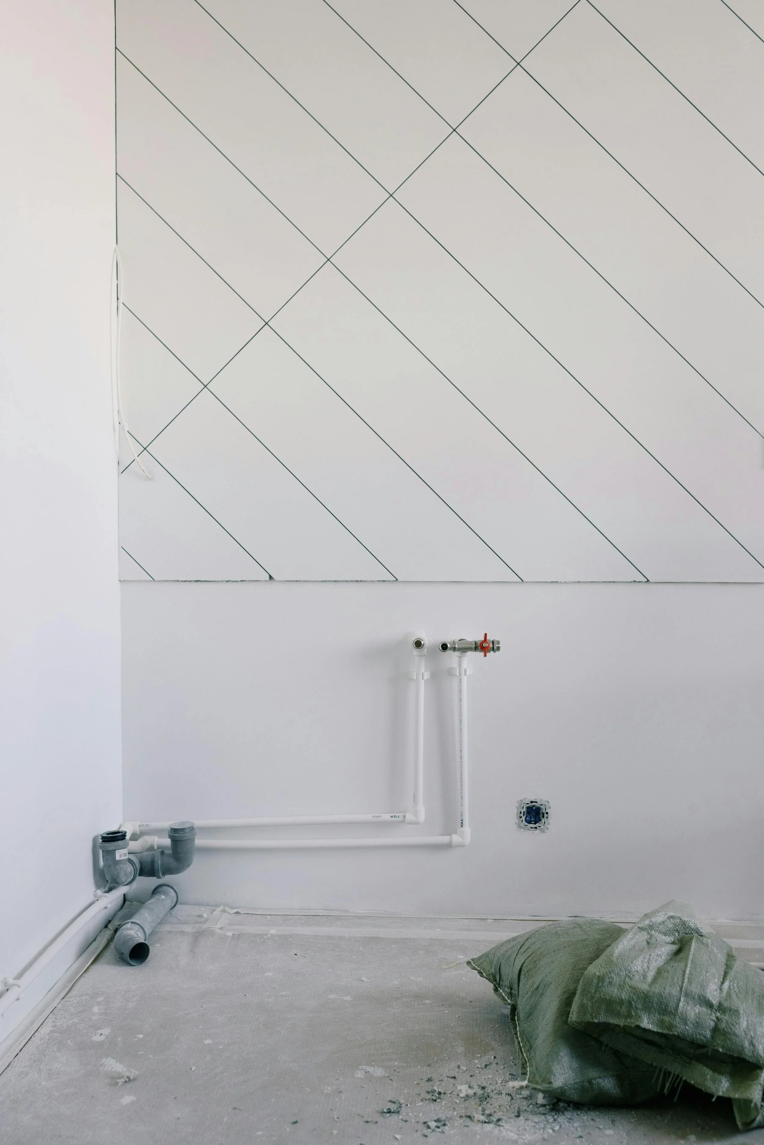 A room under construction with exposed pipes, white walls, and a section of wall with a diagonal black line pattern. There is a large black trash bag on the floor and some construction materials and debris scattered around.