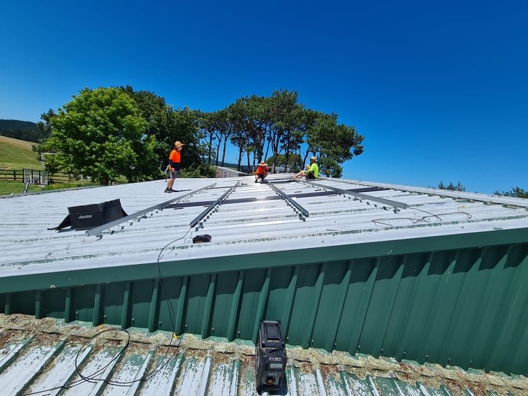 Workers installing or repairing a metal roof on a building with a green lower section on a sunny day.
