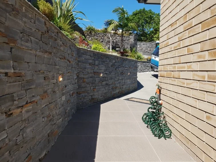A curved outdoor walkway bordered by a stone wall on the left and a brick wall on the right. The wall on the left has small lights embedded. There are garden plants and trees in the background under a clear blue sky. Several green garden hoses are coiled near the brick wall.